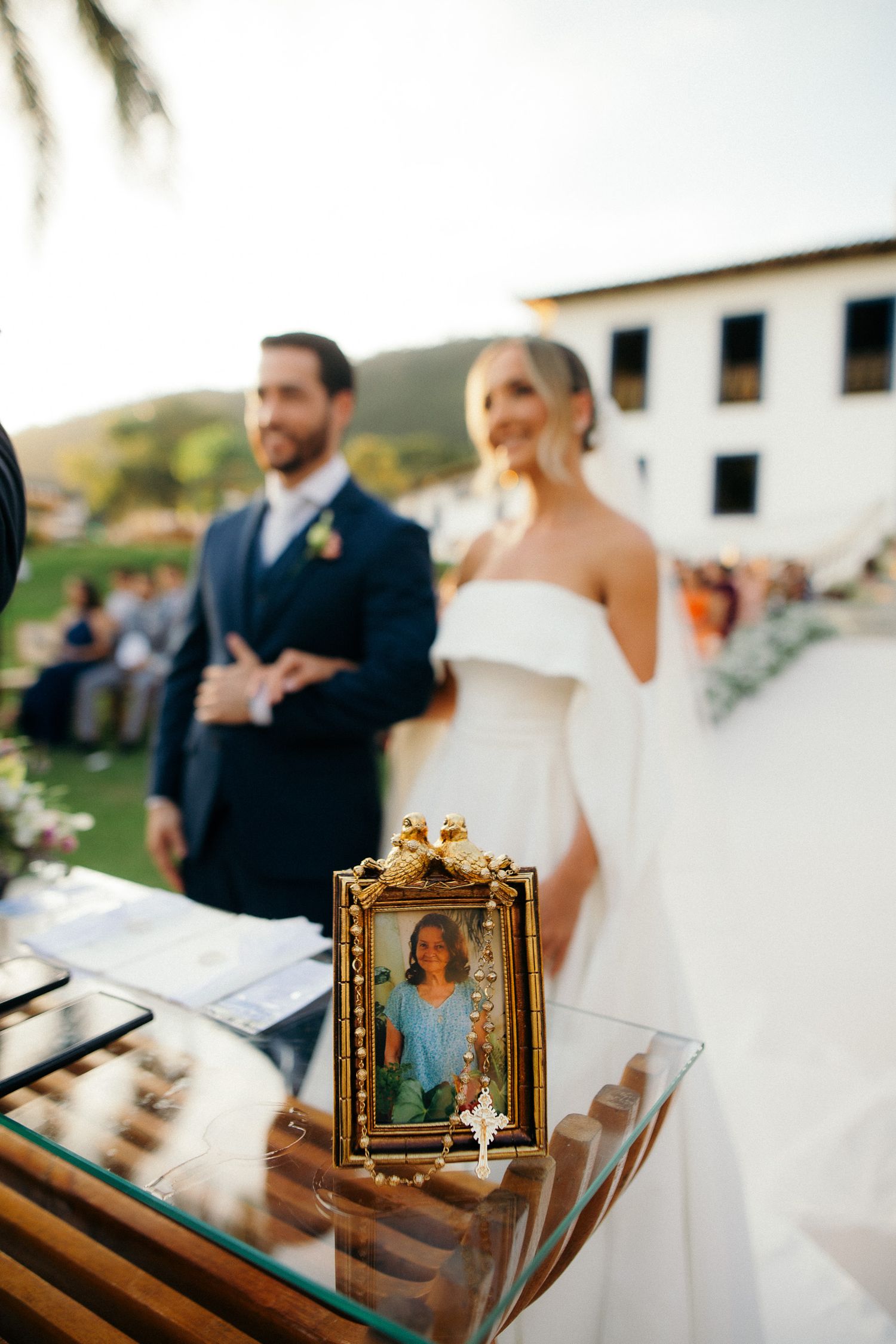 Framed memorial photo on wedding table with blurred bride and groom standing together outdoors.