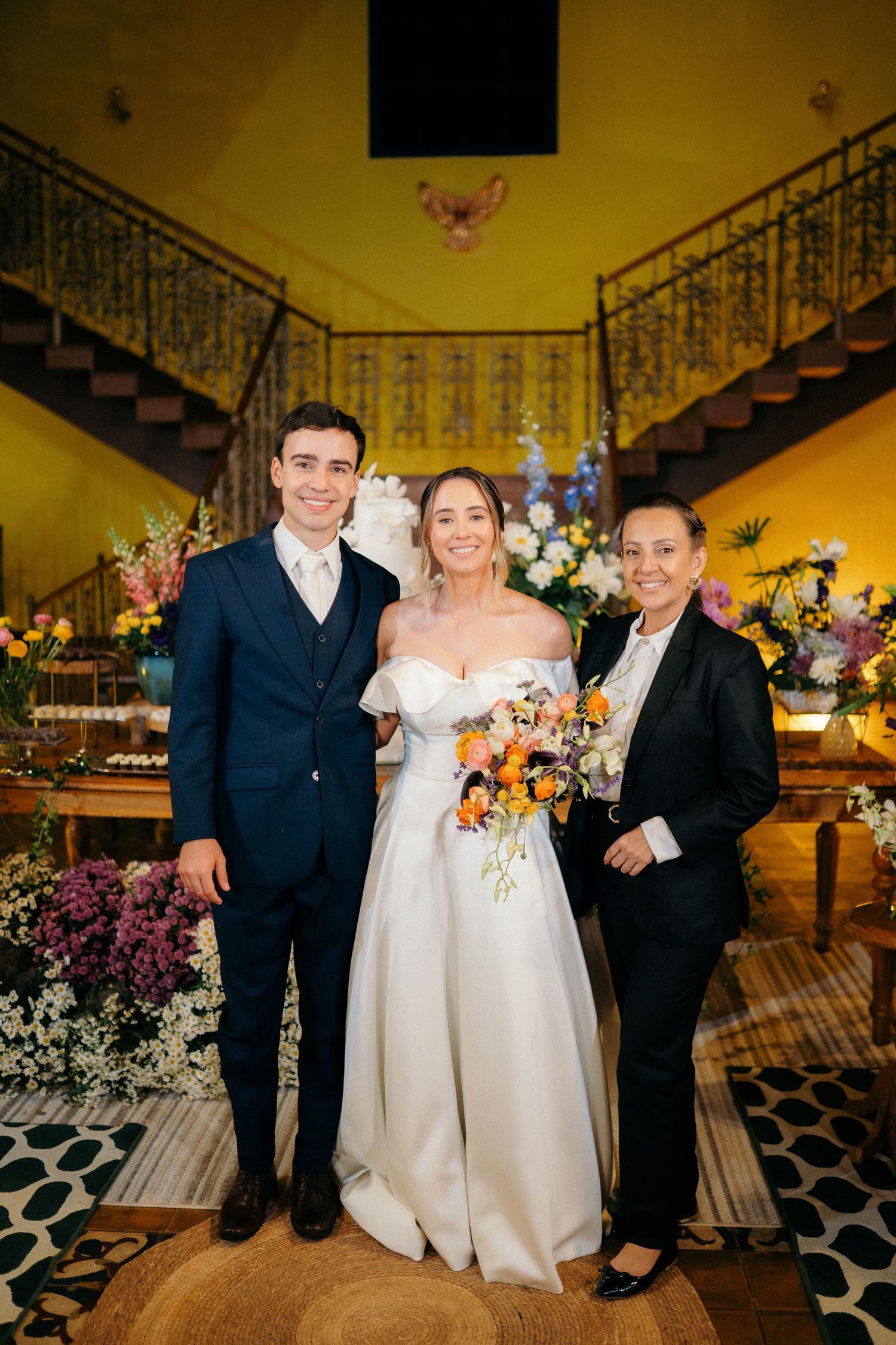 Bride in an off-shoulder gown holding a colorful bouquet poses with the groom and a guest in a decorated venue.