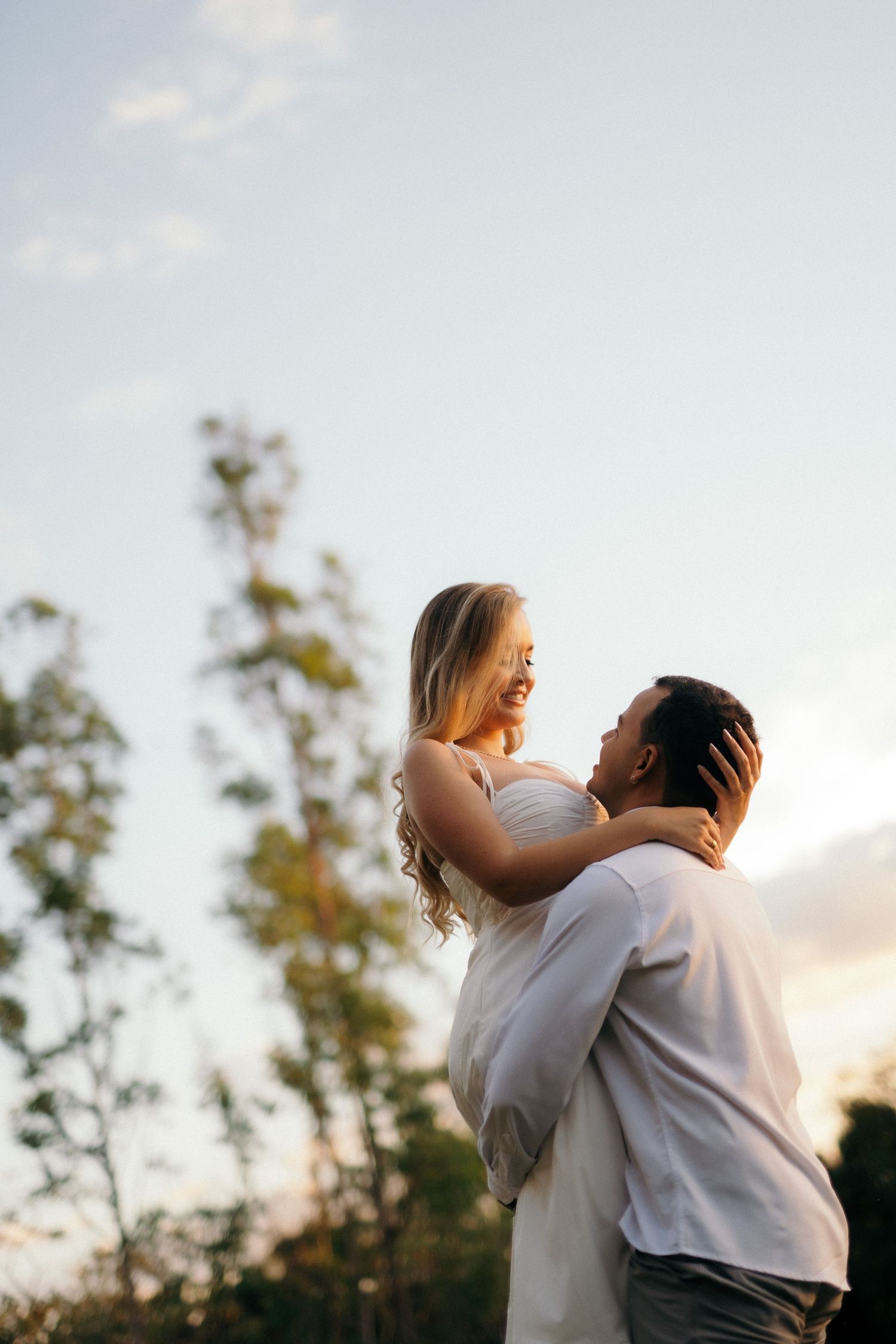 A man lifts a smiling blonde woman in a romantic outdoor sunset embrace, both dressed in white.