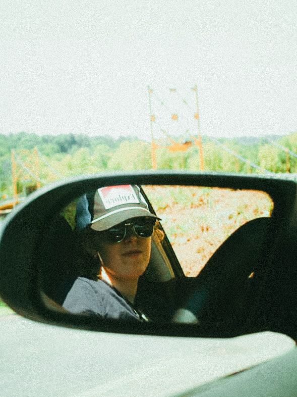 Person wearing sunglasses and a cap reflected in a car side mirror on a sunny road trip.