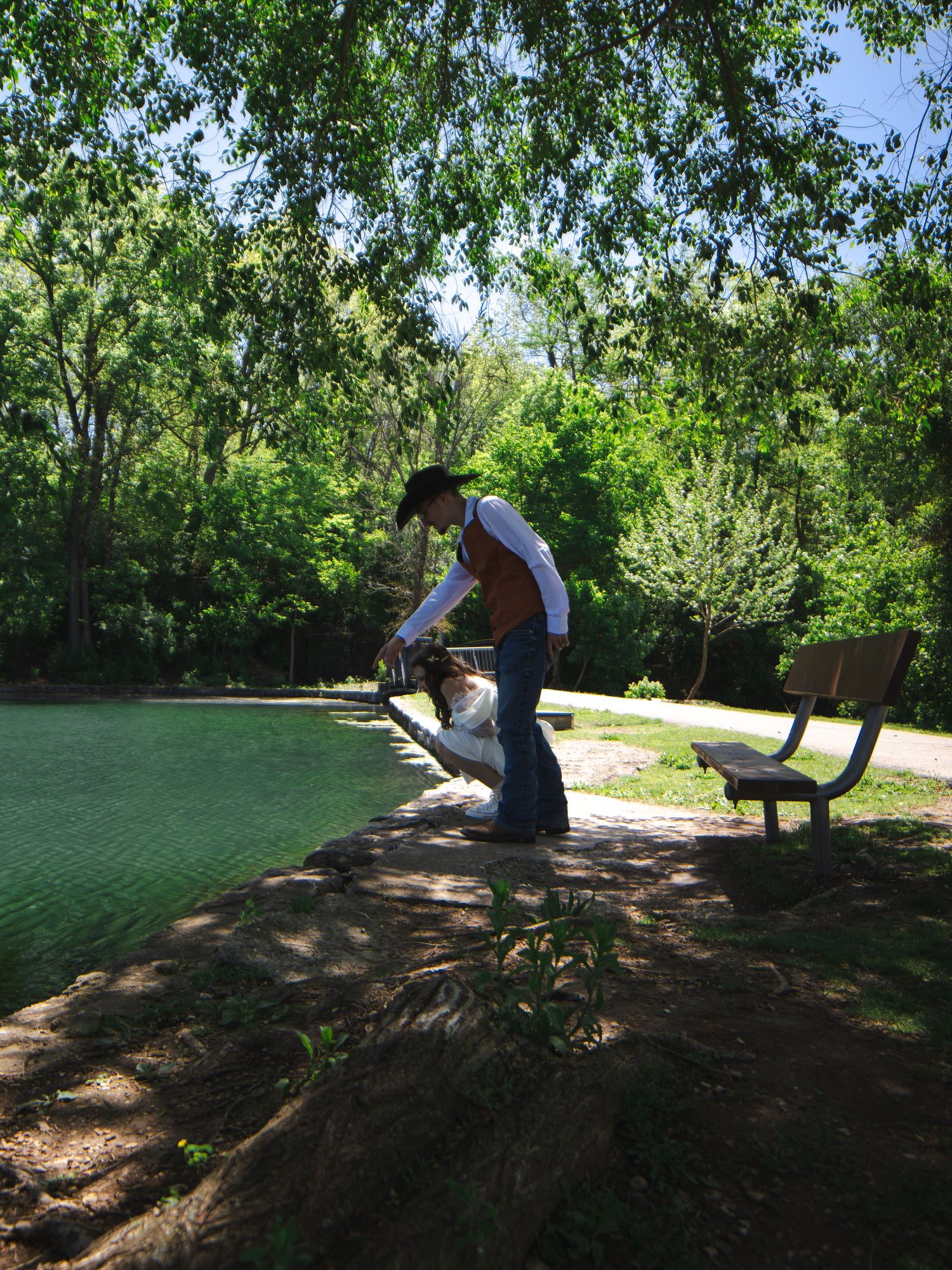 Person in cowboy hat using leaf blower near green pond with park bench and lush trees in background.