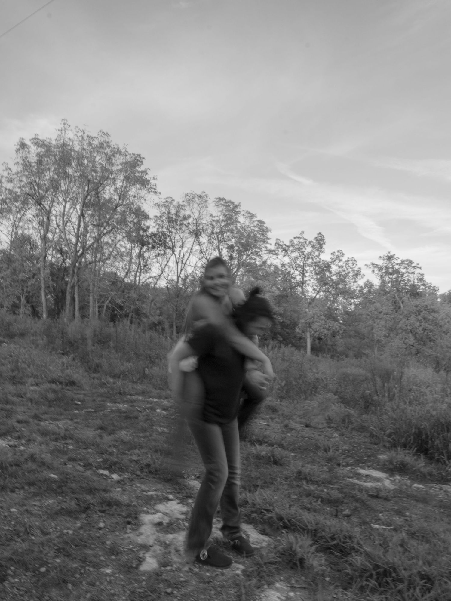 Black and white photo of two people playfully piggyback riding on a grassy hill surrounded by trees.