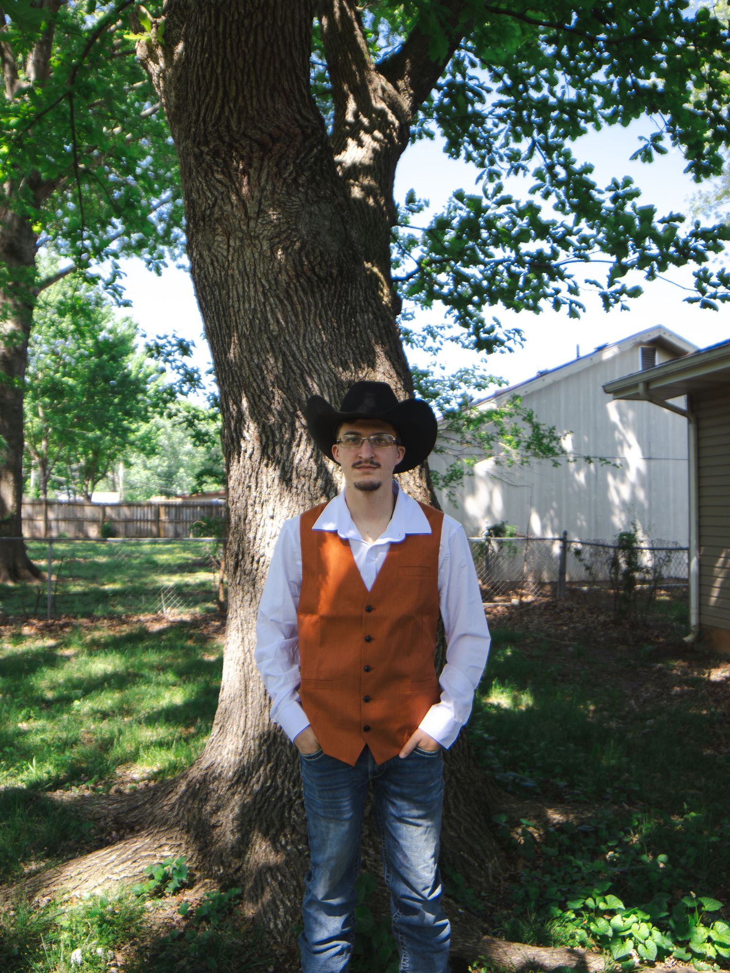 Young man in orange vest, white shirt, black cowboy hat, and jeans standing by a large tree in a green backyard.