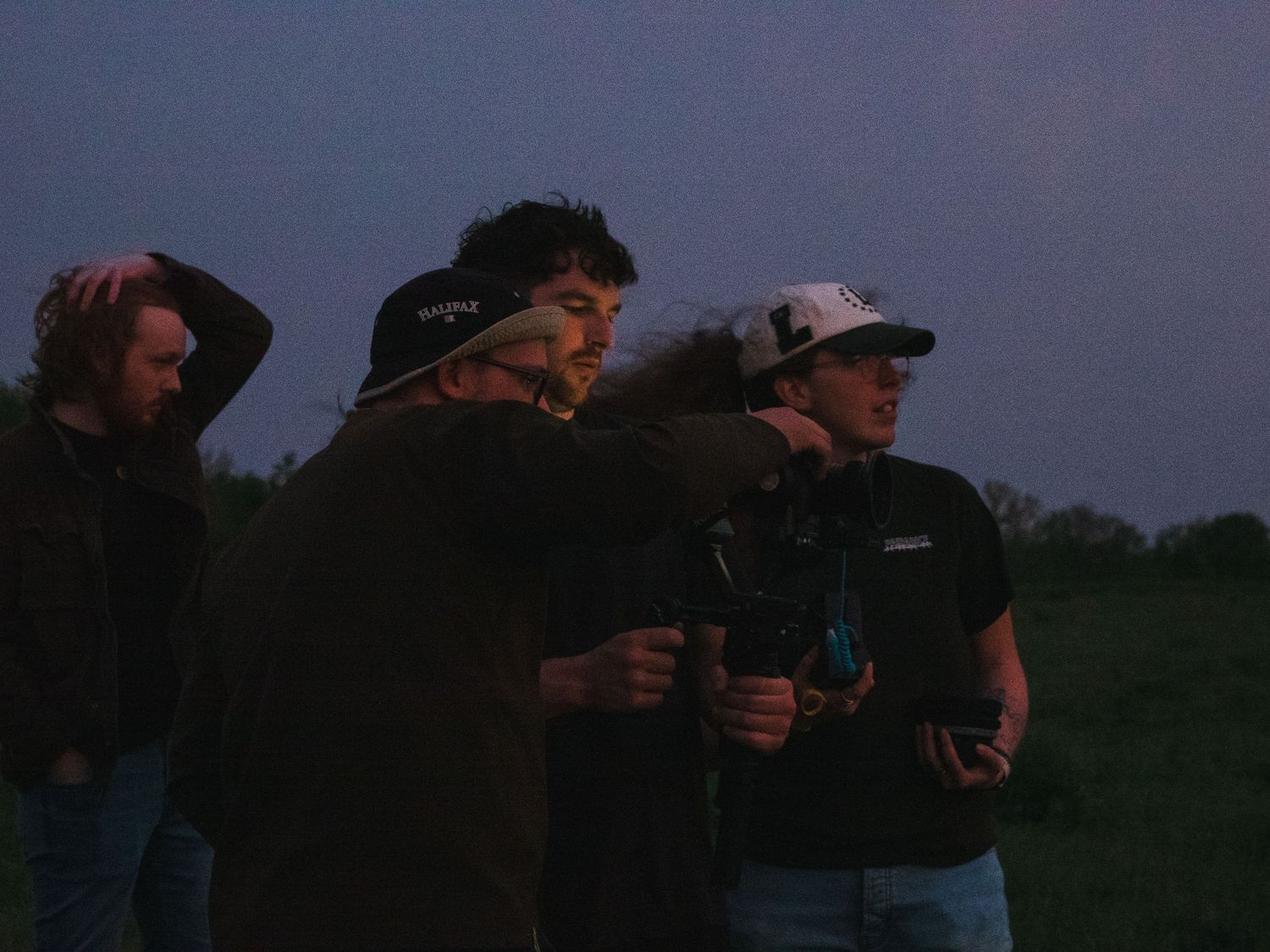 Group of young men standing outdoors at dusk, one pointing into the distance while another holds a camera.