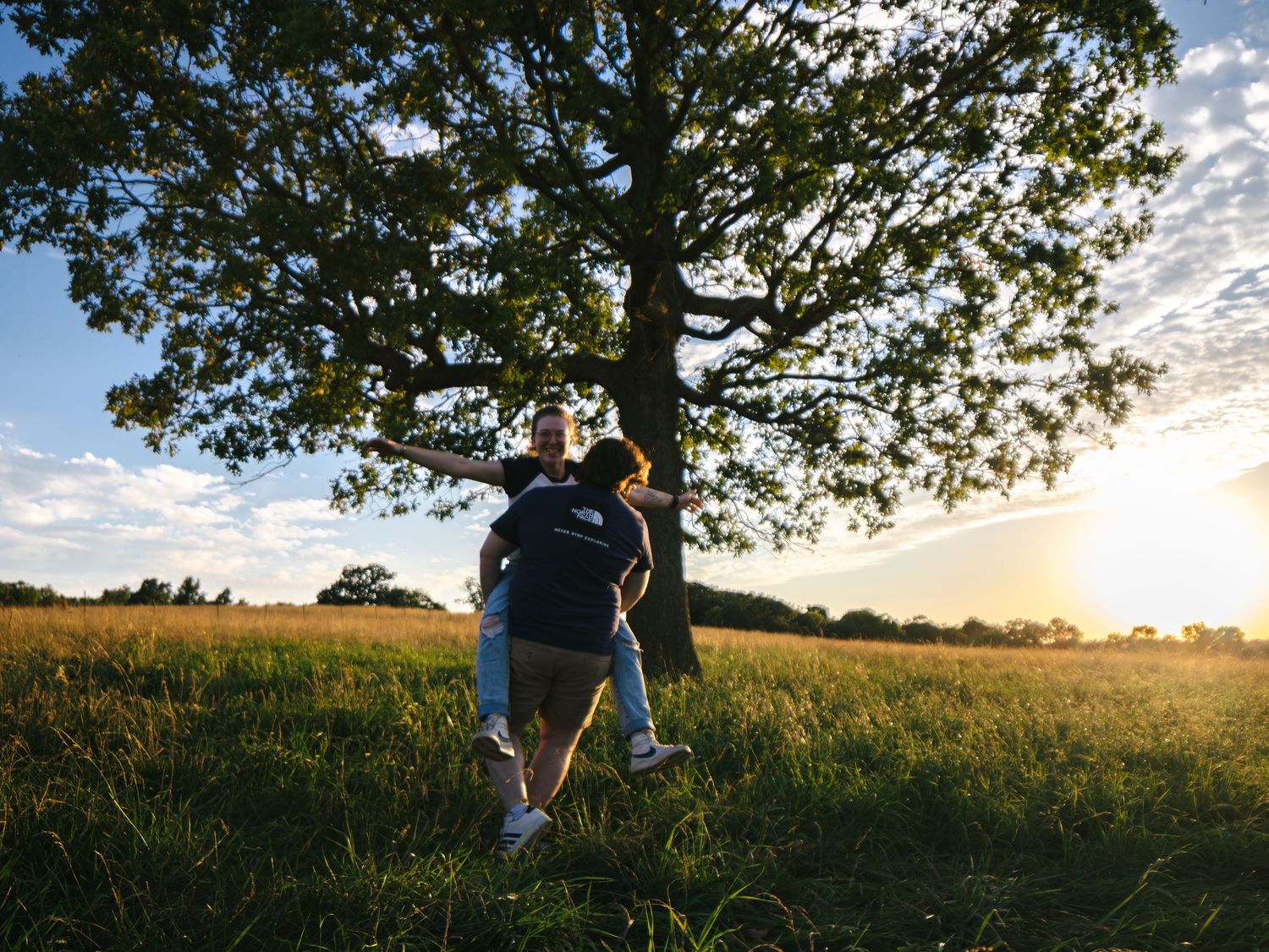 Two people embrace under a large oak tree in a sunlit green meadow at golden hour sunset.