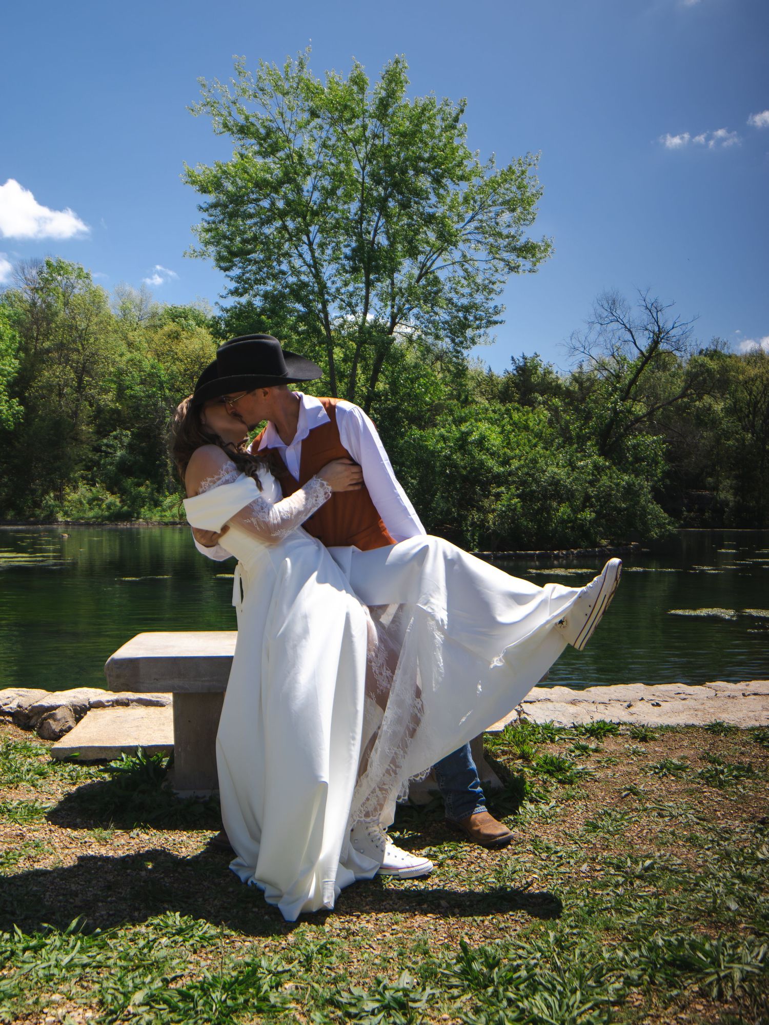 Couple sharing a romantic dip kiss in wedding attire by a serene lake, surrounded by lush green trees on a sunny day.