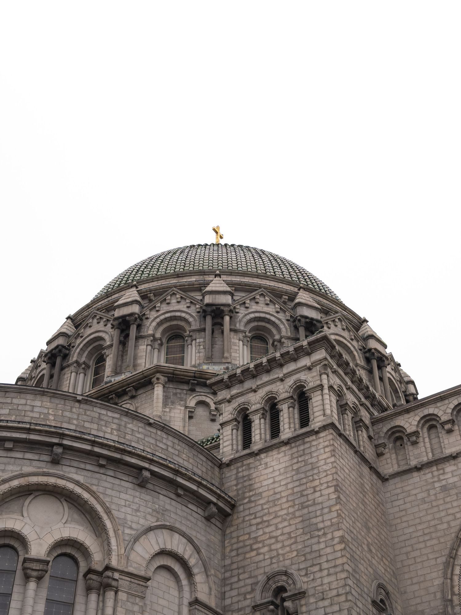 Historic stone cathedral dome with ornate arched windows and golden cross finial against overcast sky.