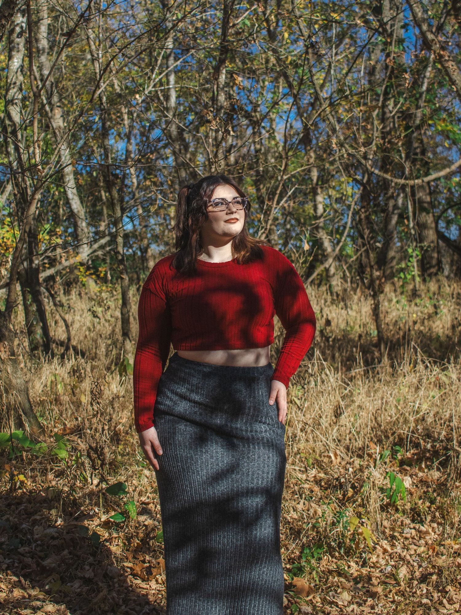 Woman in red crop sweater and gray midi skirt standing outdoors among autumn trees on a sunny day.