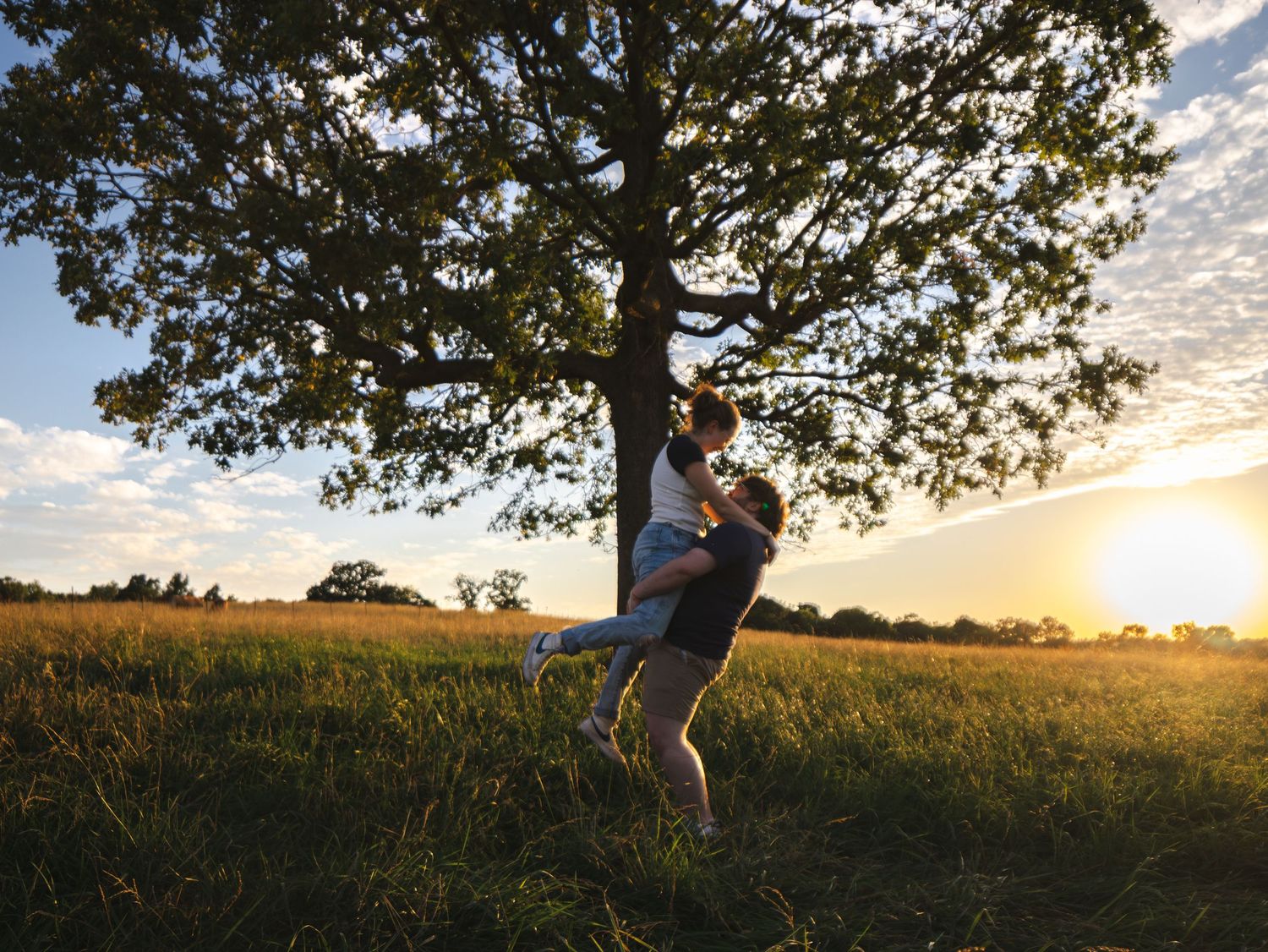 Couple embracing romantically under a large oak tree in a golden sunset meadow.