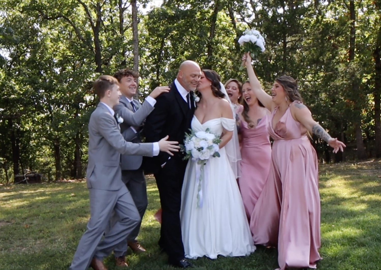 Wedding couple kissing surrounded by cheering bridesmaids in pink gowns and groomsmen in gray suits outdoors.