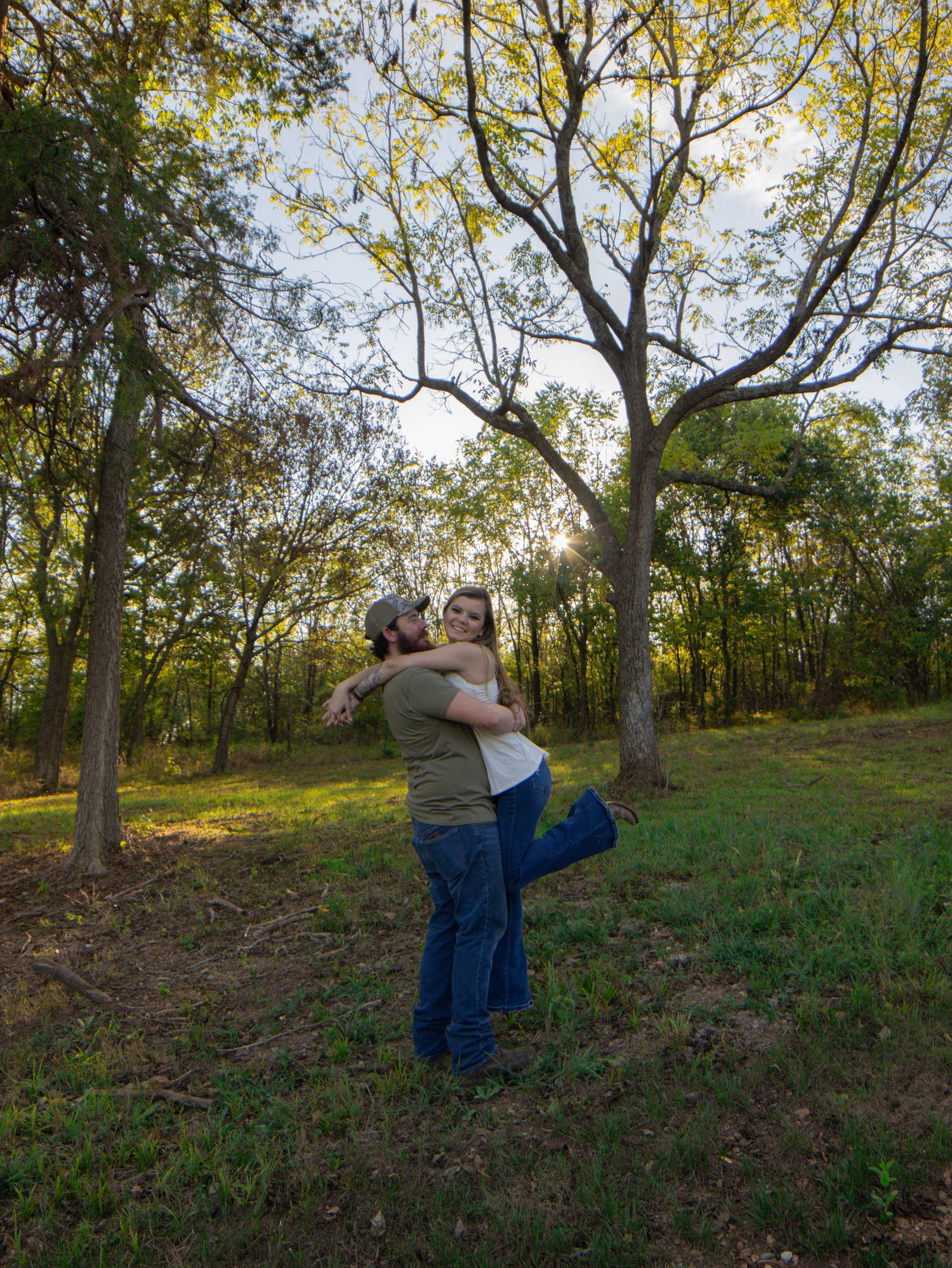 Couple embracing and spinning in a sunlit forest clearing surrounded by lush green trees.