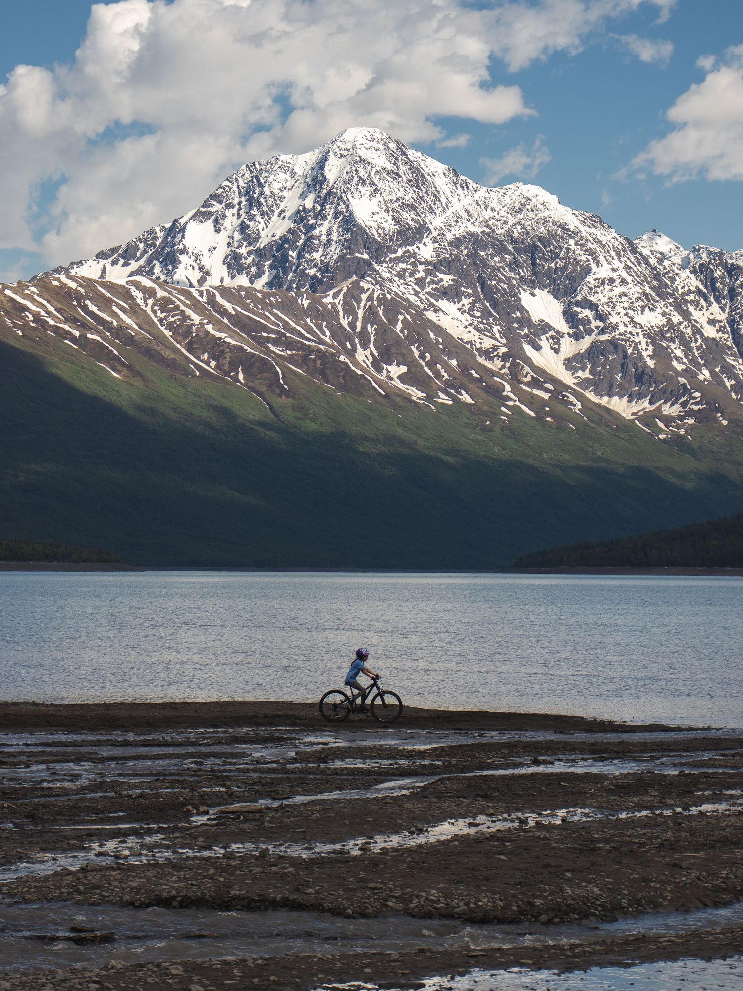 Cyclist riding along a lakeside trail with a snow-capped mountain backdrop under a partly cloudy sky.
