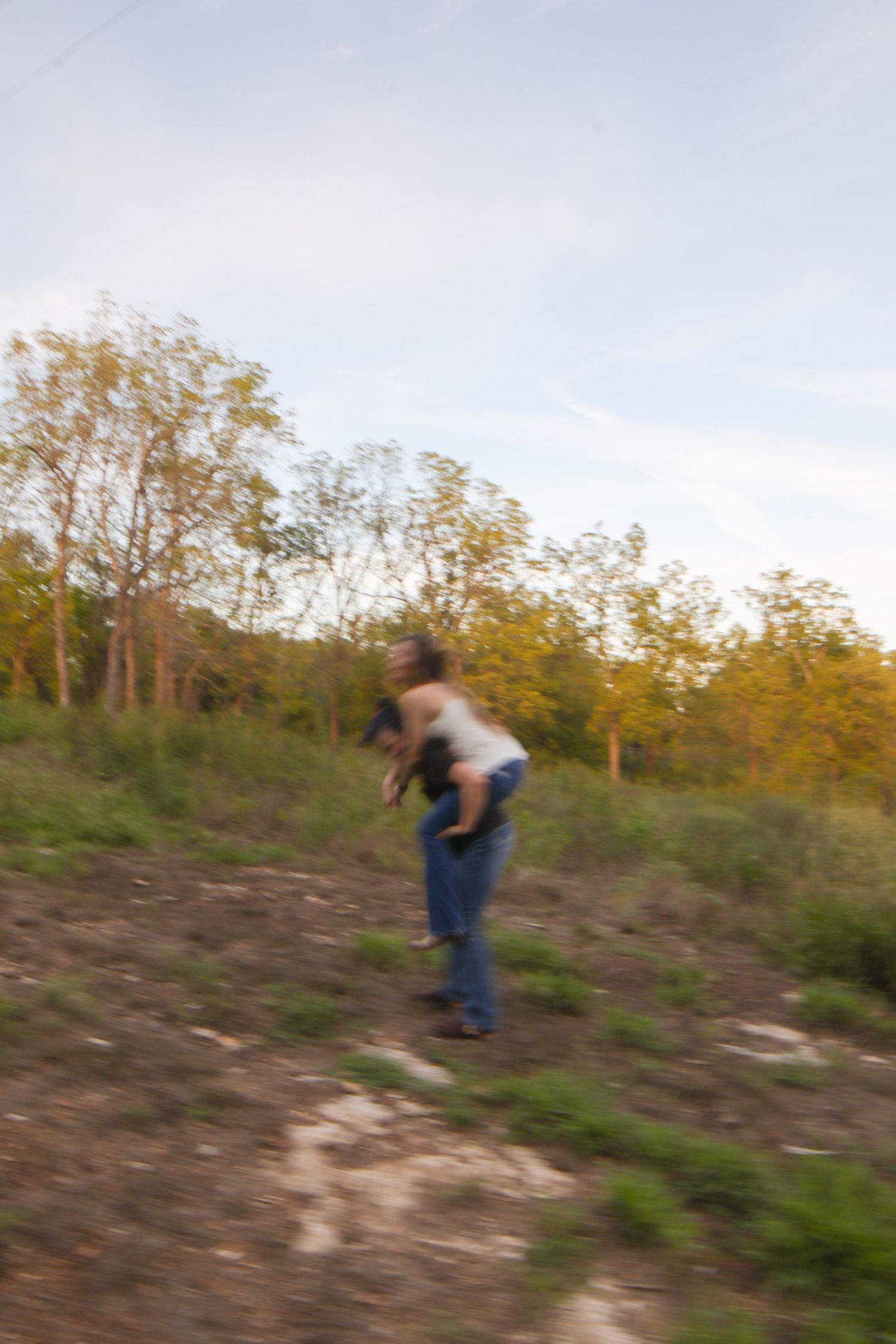 A person carrying someone on their back outdoors on a hillside surrounded by autumn trees under a cloudy sky.