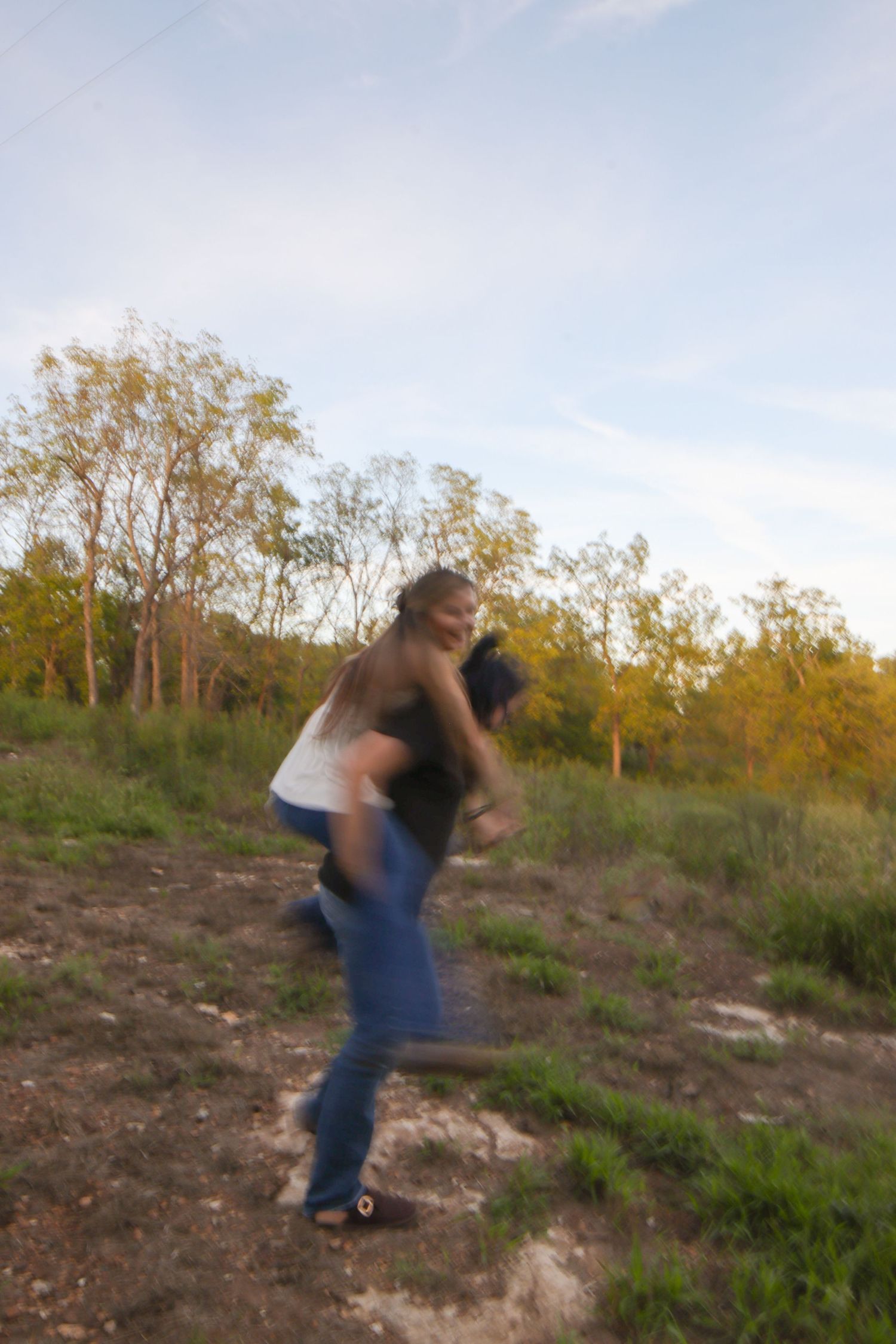 Two people playfully spinning outdoors on a grassy hillside with trees in the background during golden hour.