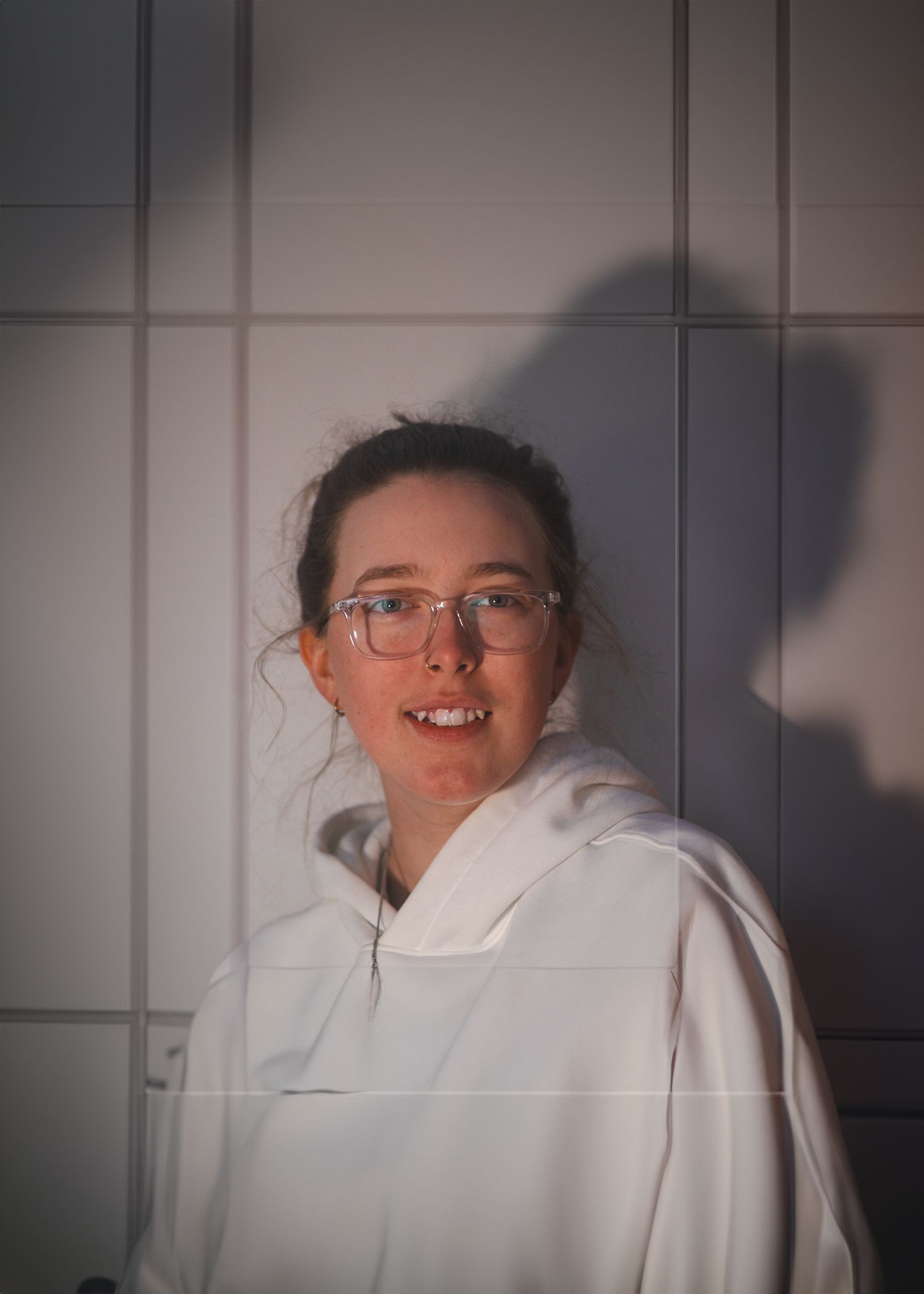 Young woman with glasses smiling in white hoodie against tiled wall with dramatic shadow lighting.