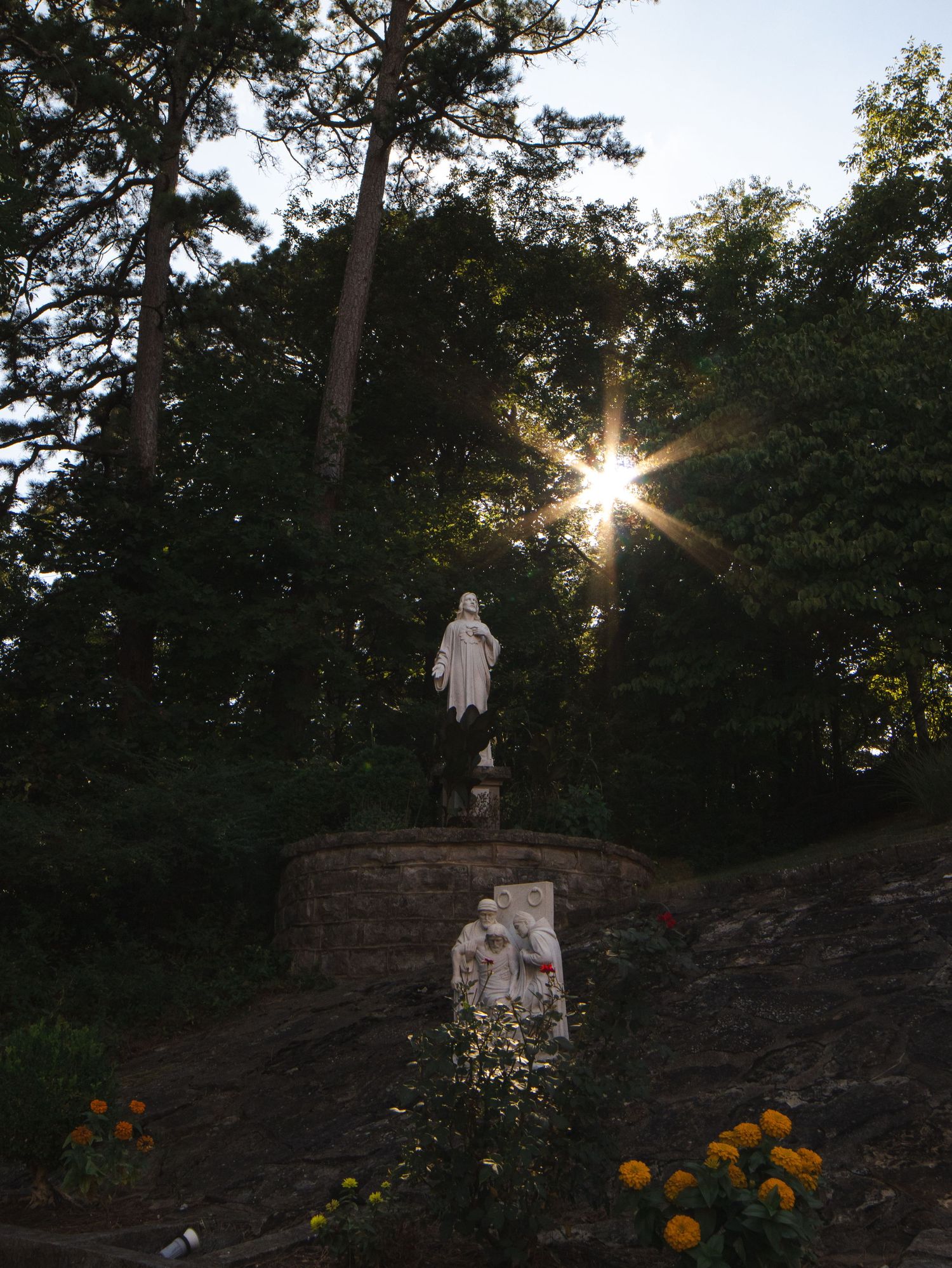 White religious statue on stone pedestal surrounded by trees with sunburst effect and yellow flowers in foreground.