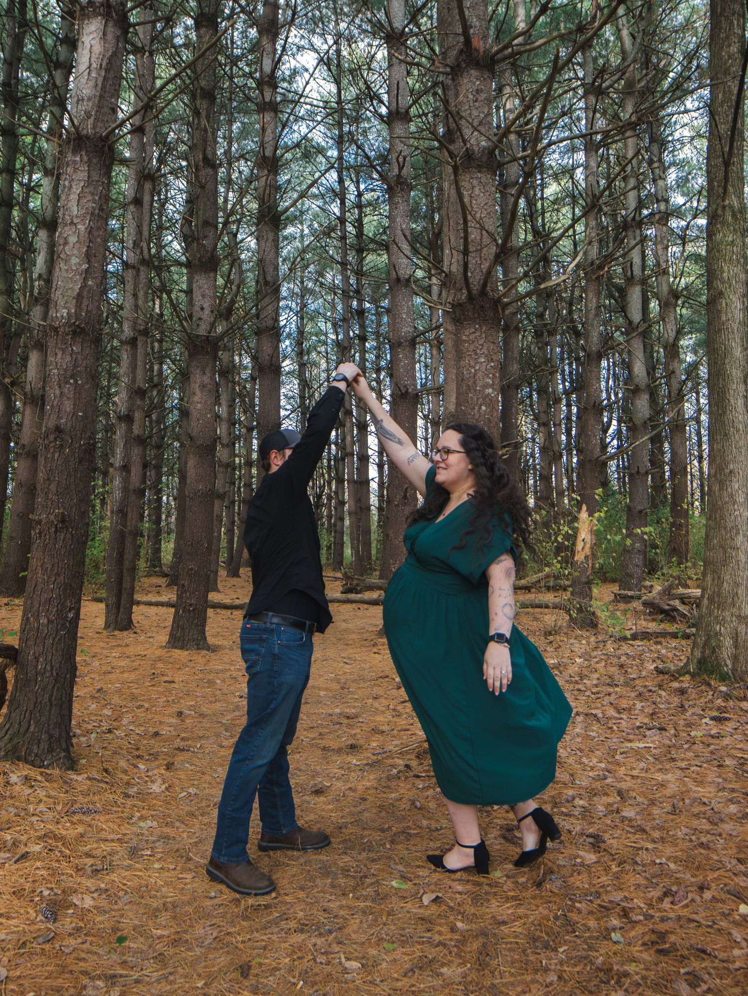 Couple dancing together in a pine forest, woman in green dress and man in black shirt spinning outdoors.