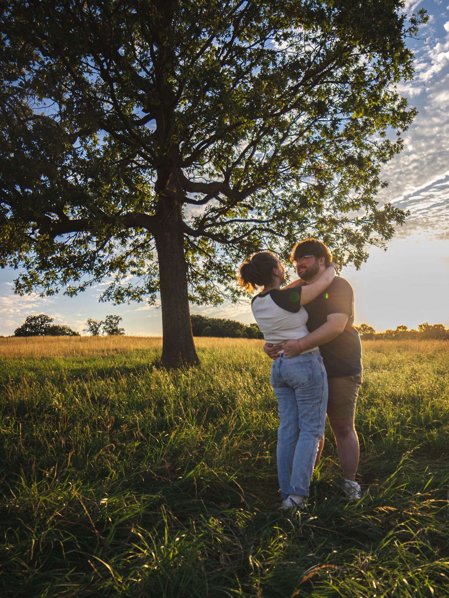 Couple embracing romantically under a large oak tree in a sunlit green meadow at golden hour.
