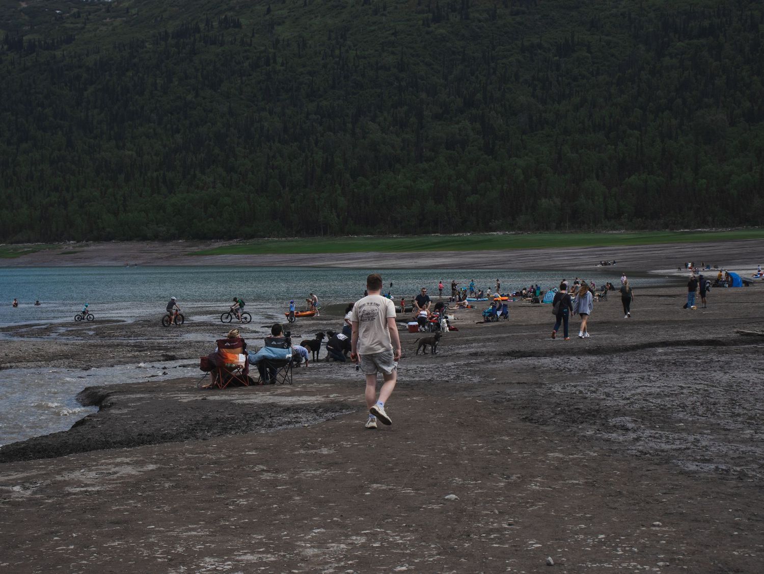 People gathering on a rocky shoreline beside a glacial blue lake surrounded by lush green forested mountains.