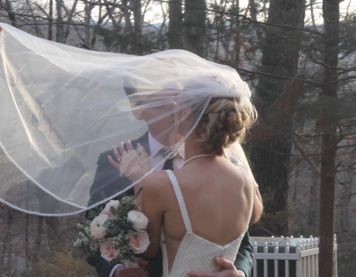 Bride and groom sharing a romantic kiss outdoors, with wedding veil blowing in the wind, bride holding a floral bouquet.