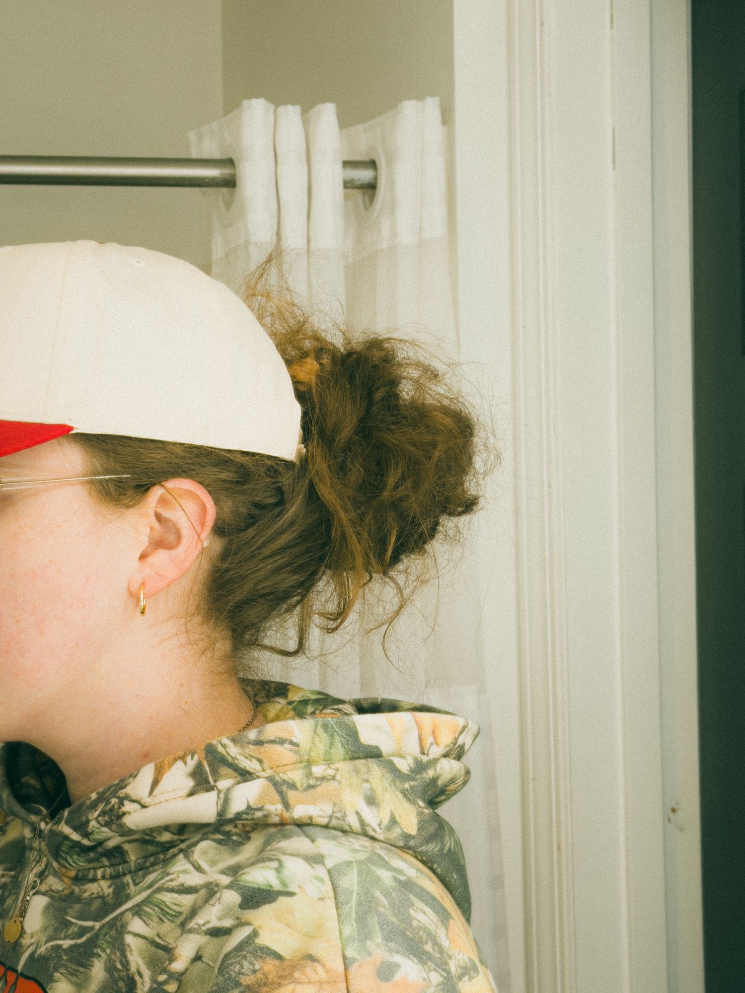 Person wearing camo hoodie and white snapback cap with hair in messy bun, standing near curtained window.