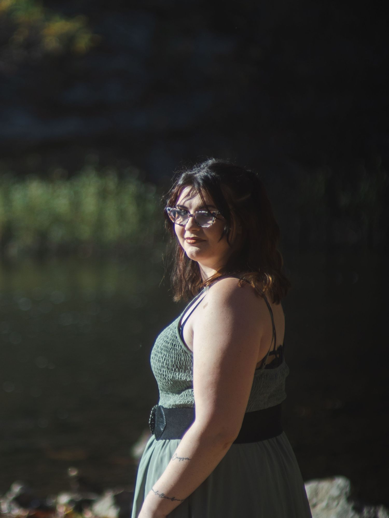 Young woman in green sundress and cat-eye sunglasses poses outdoors near a sunlit rocky waterside.