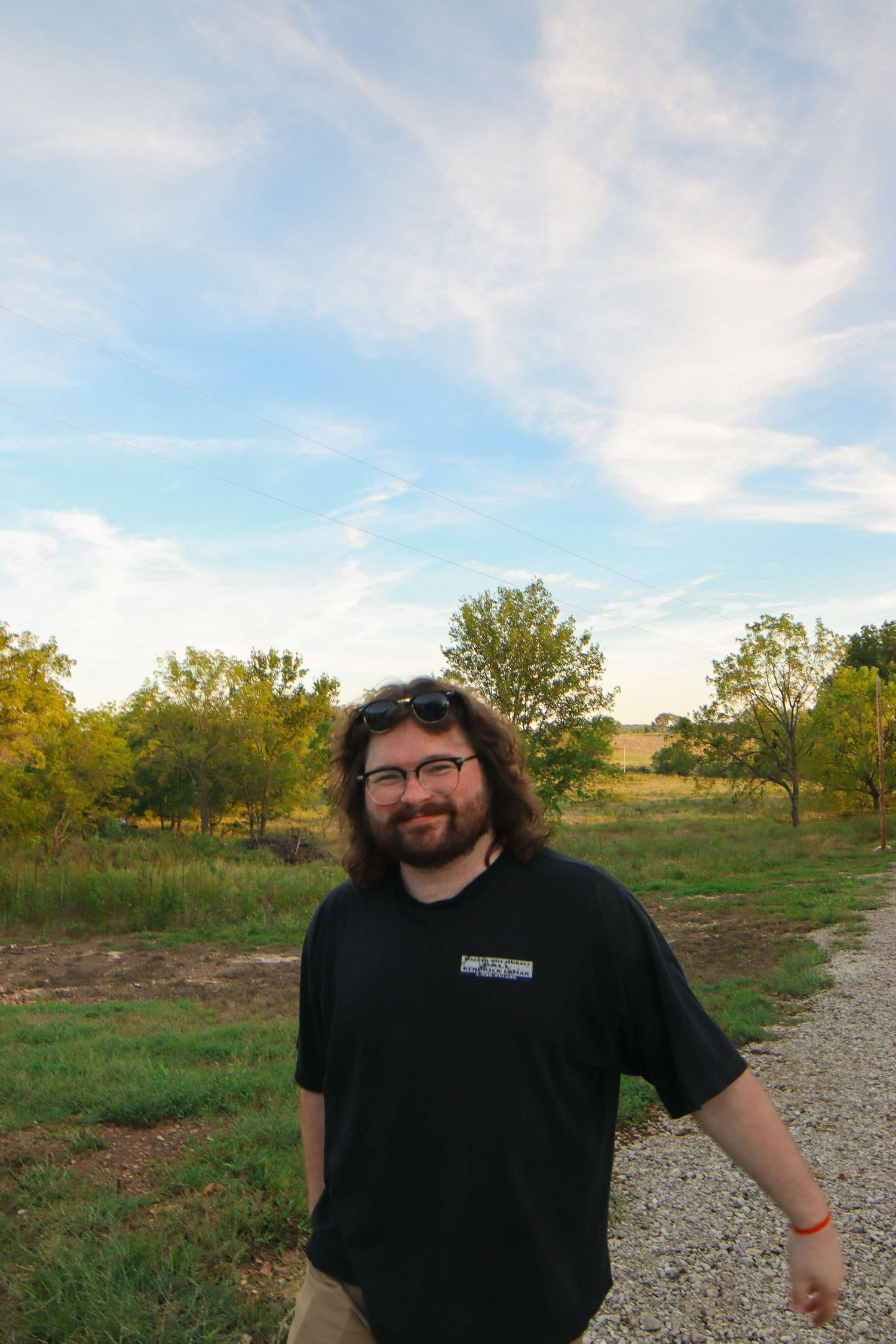 Smiling man with beard and glasses in black t-shirt standing on gravel path surrounded by green trees and open sky.
