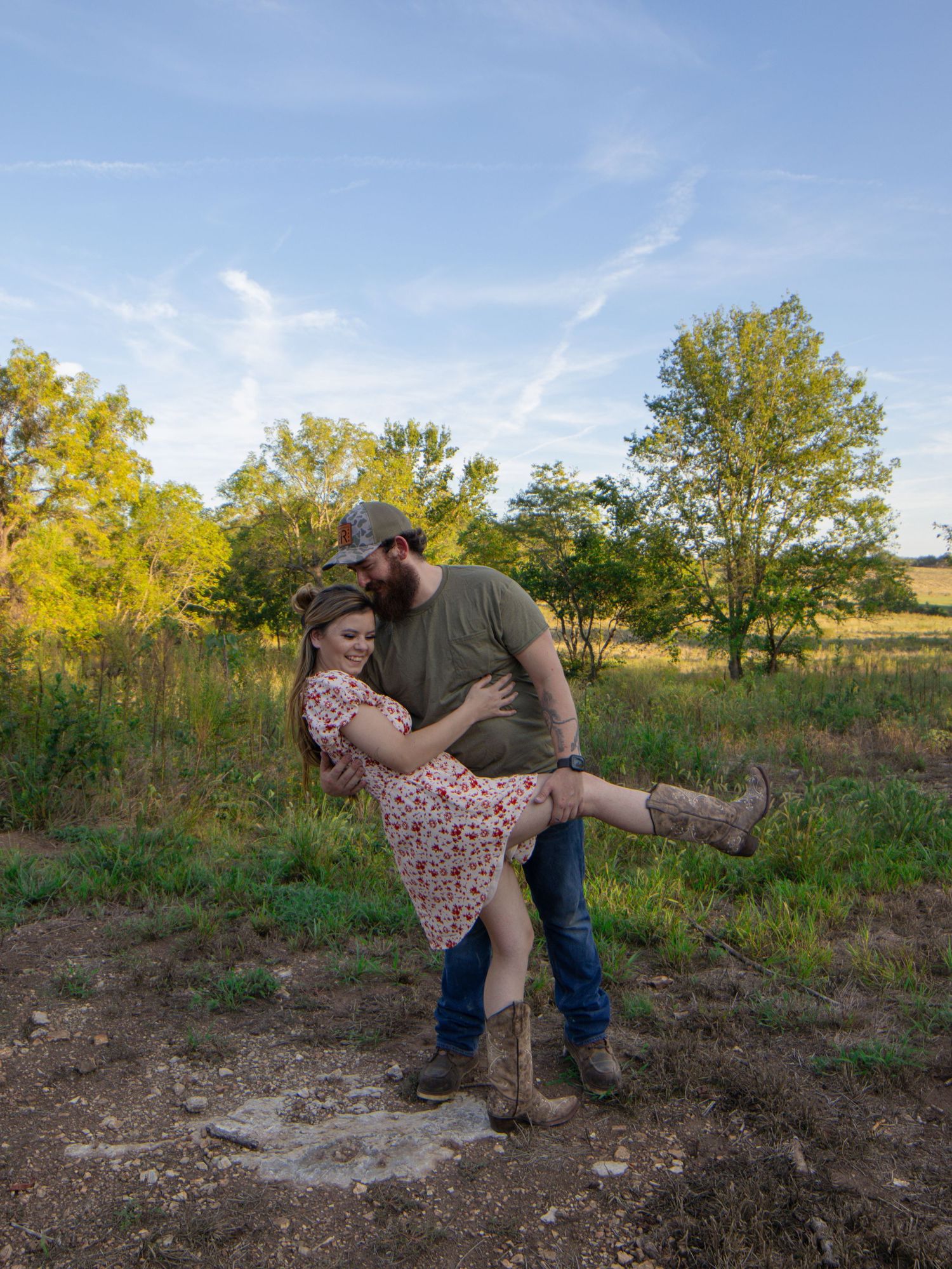 Couple embracing outdoors in a field, man lifting woman in floral dress and cowboy boots, green trees and blue sky behind them.