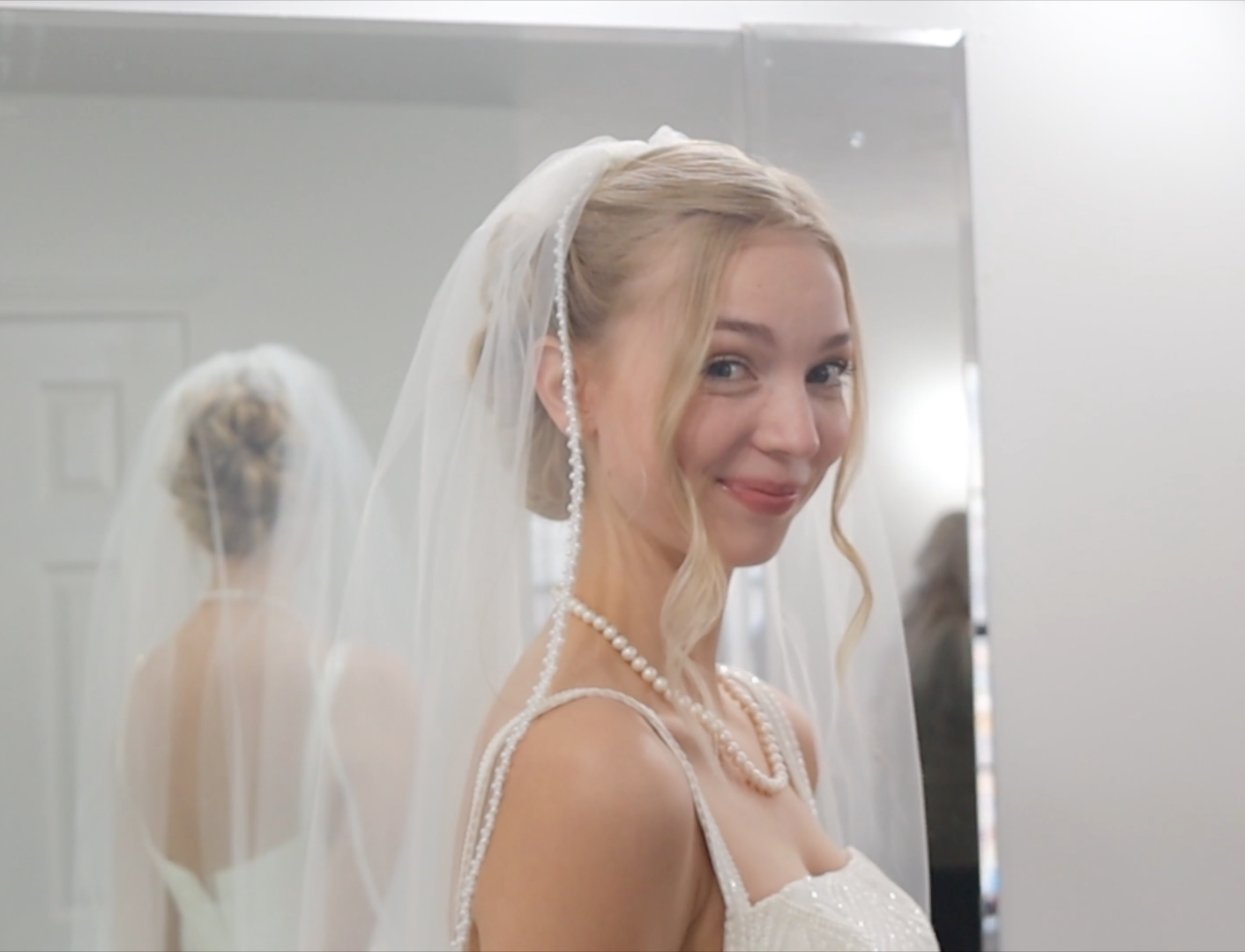 Bride in white gown and veil wearing pearl necklace smiling over shoulder, reflected in mirror.