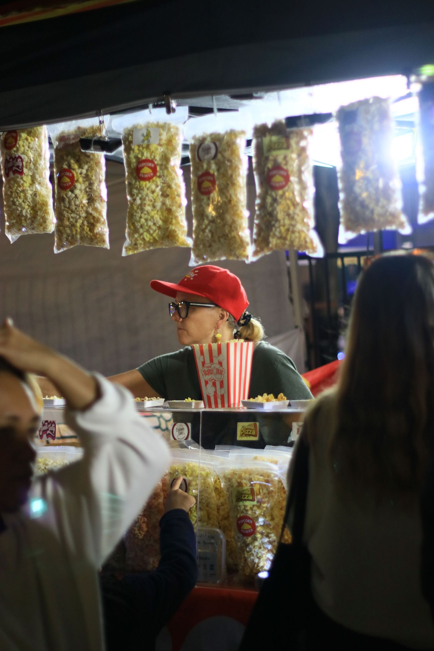 A vendor in a red cap and sunglasses serving popcorn at a market stall with bags of popcorn hanging overhead.