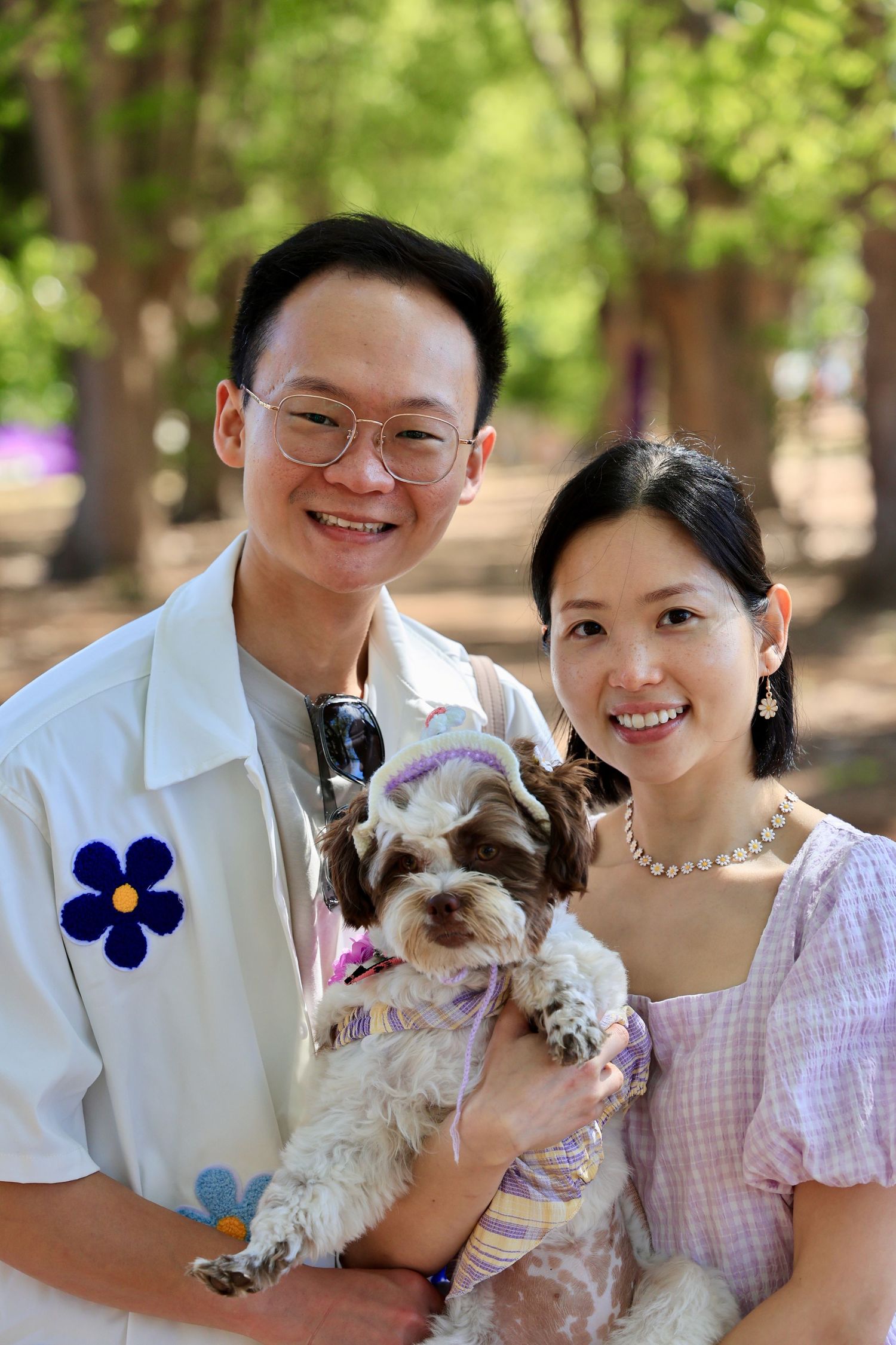 Smiling Asian couple holding an adorable dressed-up Shih Tzu dog outdoors in a lush green park setting.