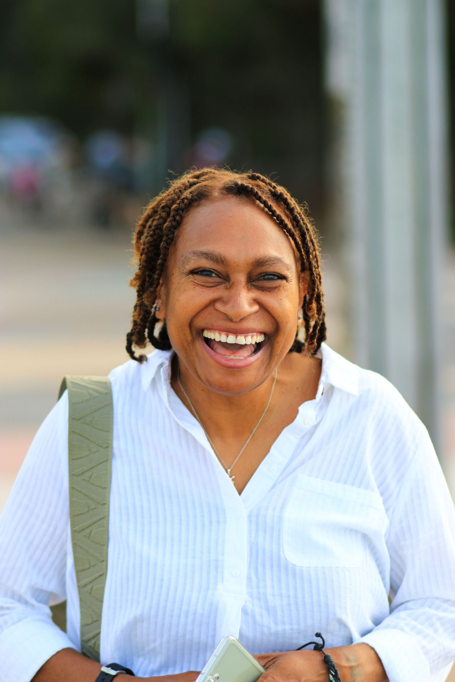 A joyful Black woman with braided hair laughing outdoors, wearing a white striped shirt.