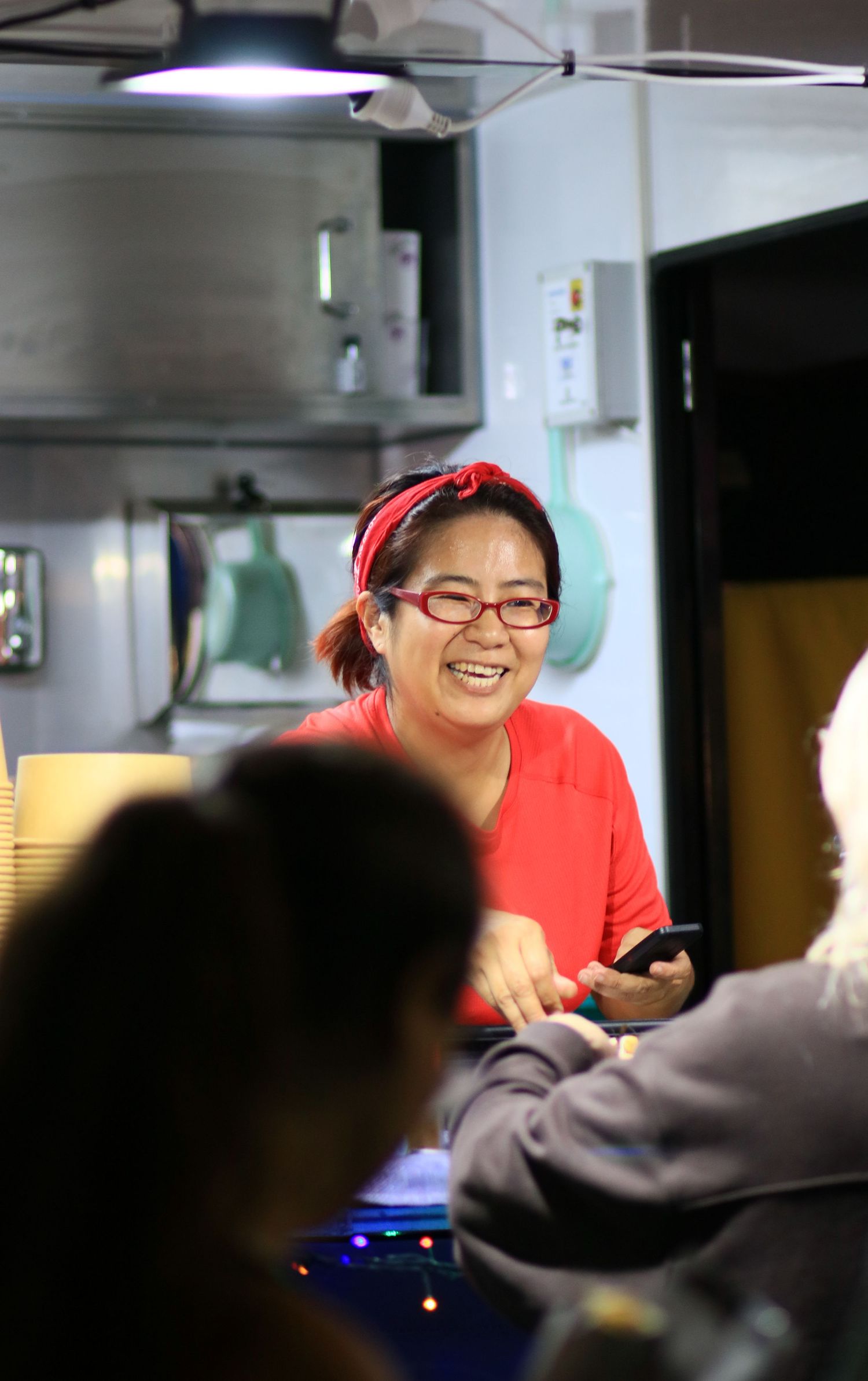 A smiling Asian woman wearing red glasses and a red headband working behind a counter in a kitchen.