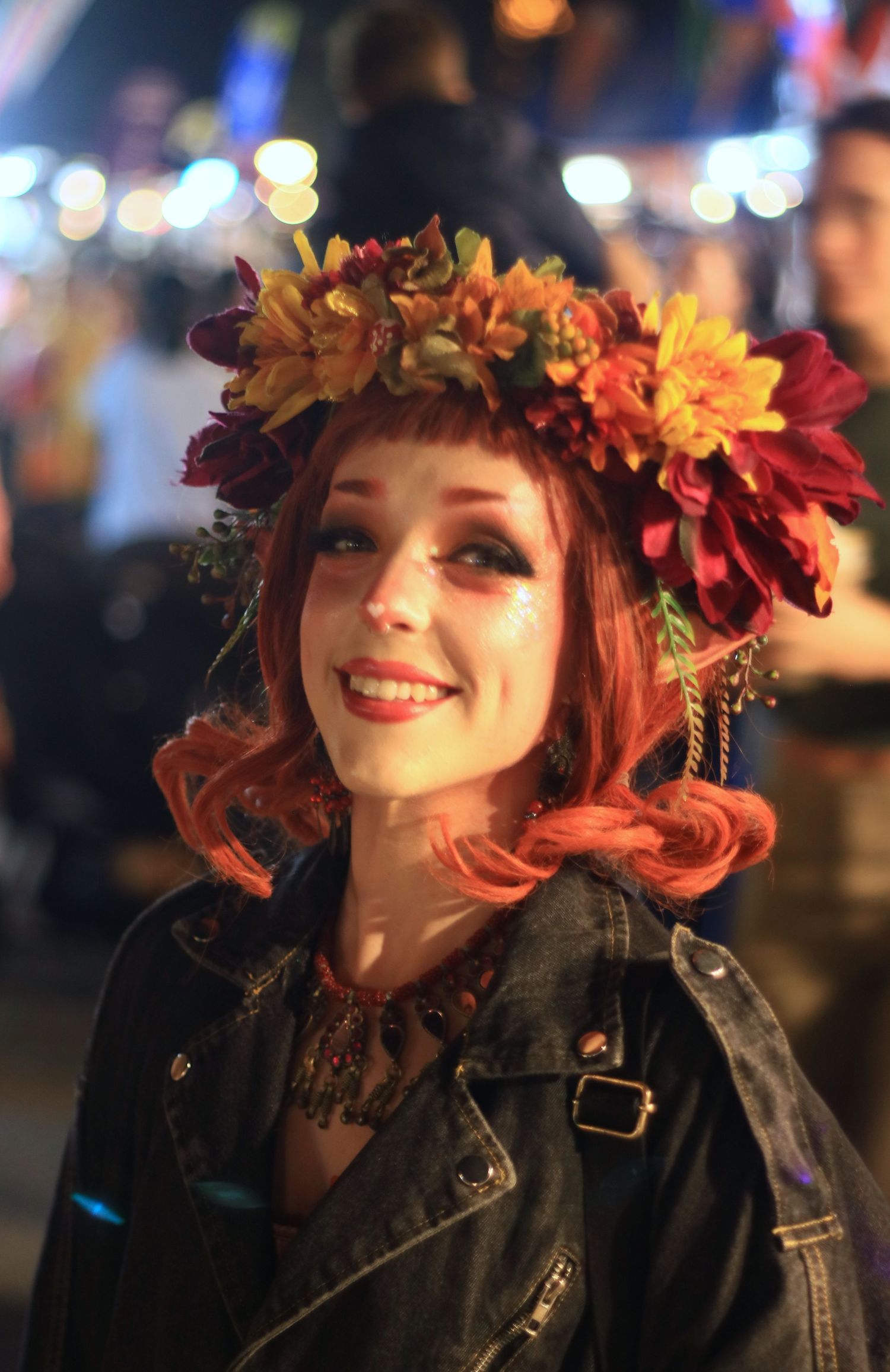 Young woman with red hair wearing autumn flower crown and leather jacket smiling at outdoor night event.