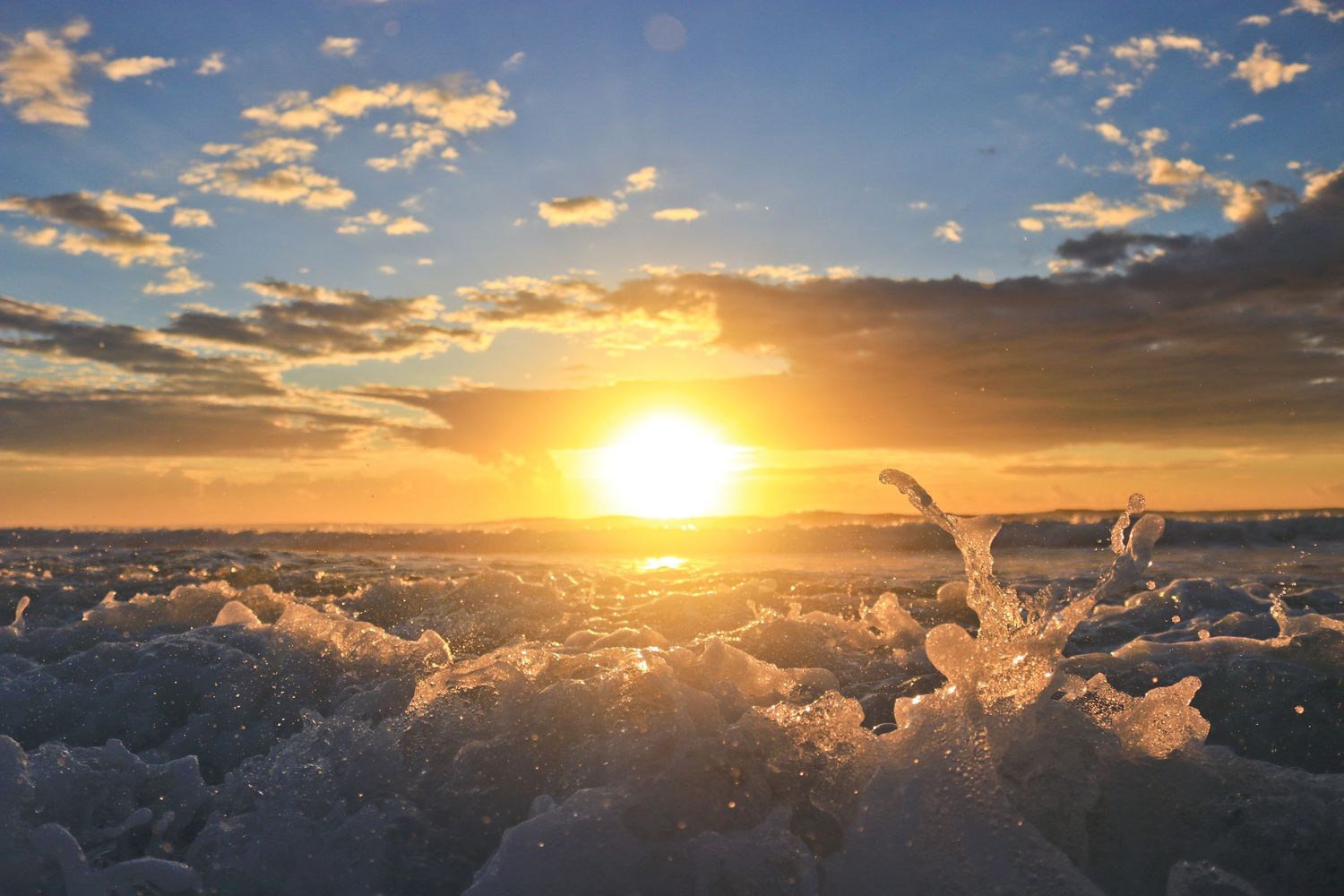 Dramatic ocean waves crashing at golden sunset with glowing sky and scattered clouds above.
