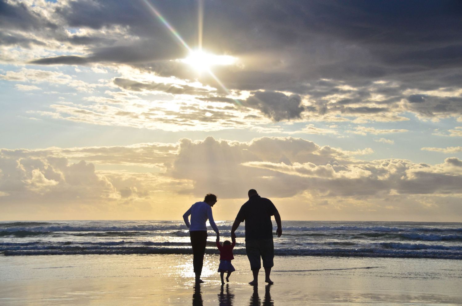 Two adults and a child silhouetted against a dramatic sunset sky while walking on a beach together.