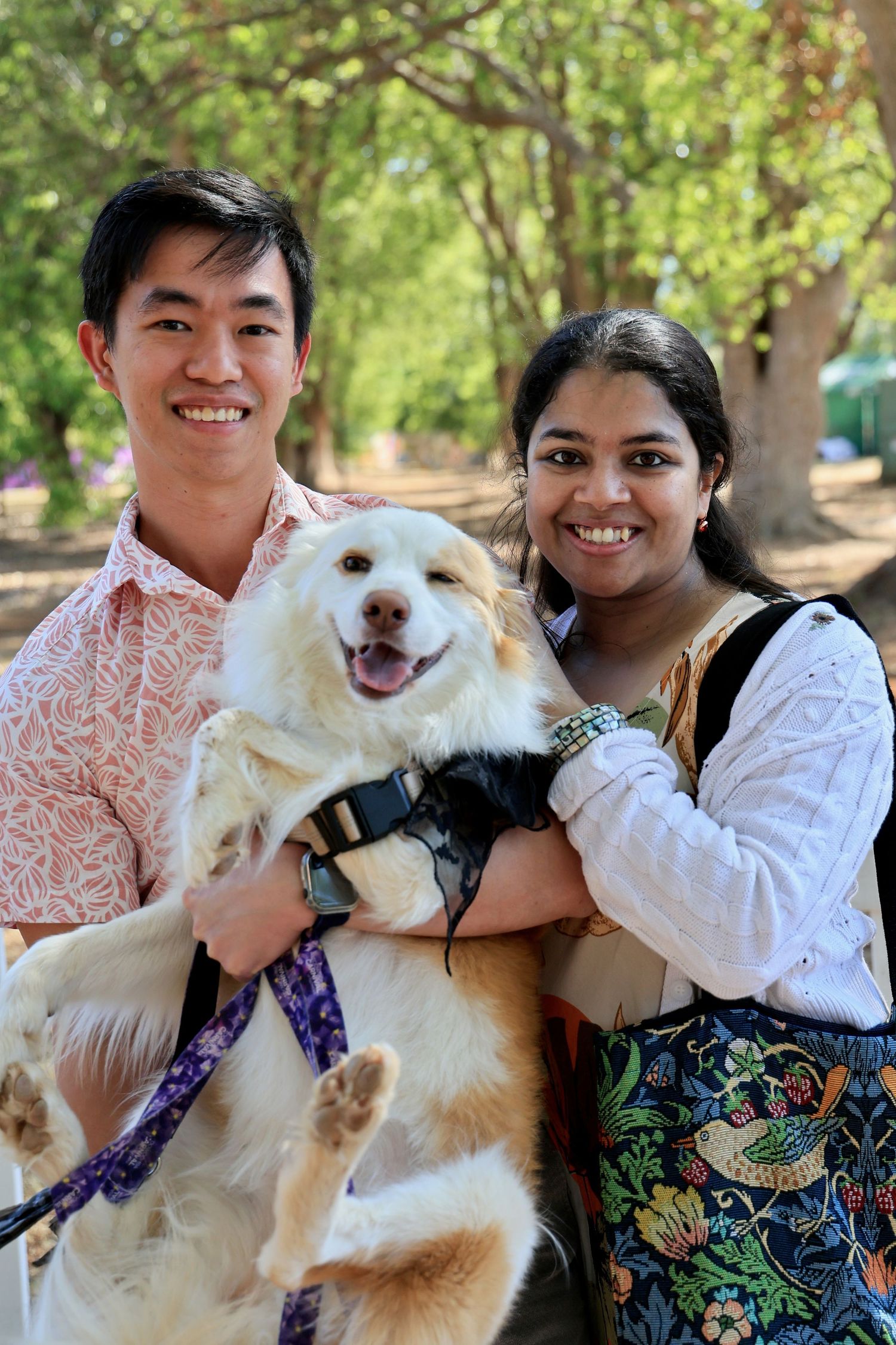 Two smiling people holding a happy fluffy dog outdoors in a tree-lined park setting.