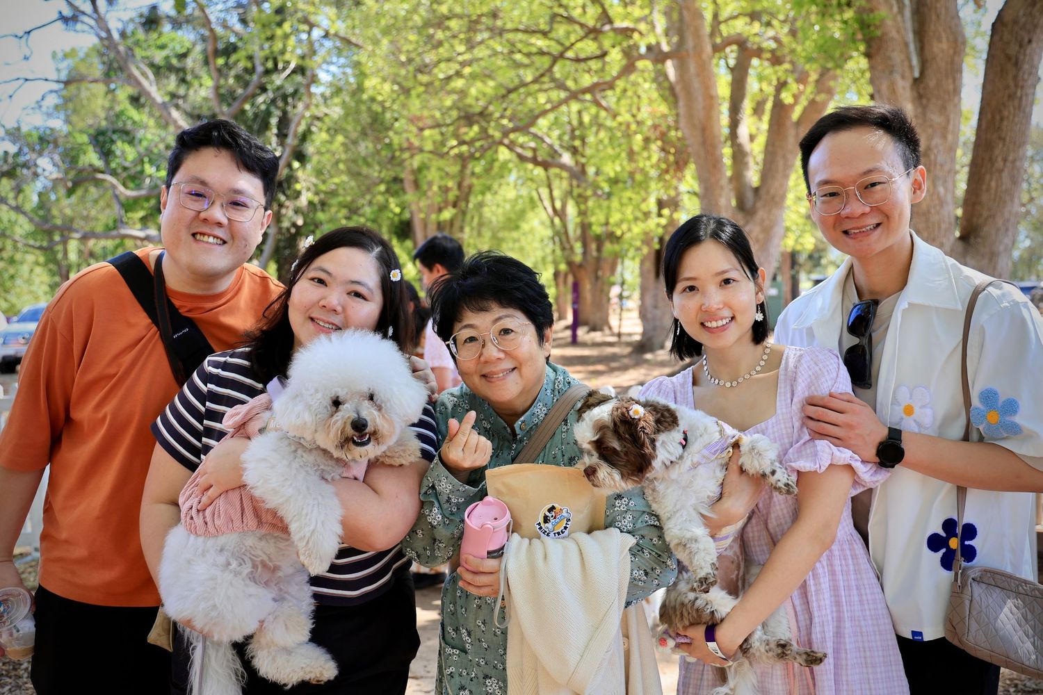 Happy family group of five people posing outdoors with two fluffy dogs, smiling together under shady trees.