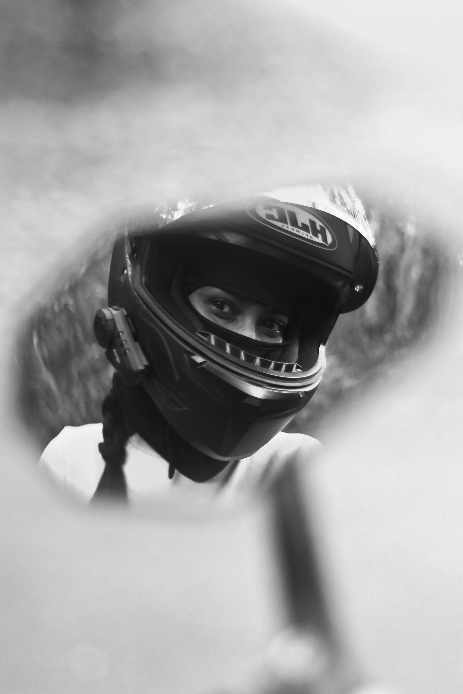 A black and white reflection of a motorcycle rider wearing a full-face HJC helmet in a side mirror.