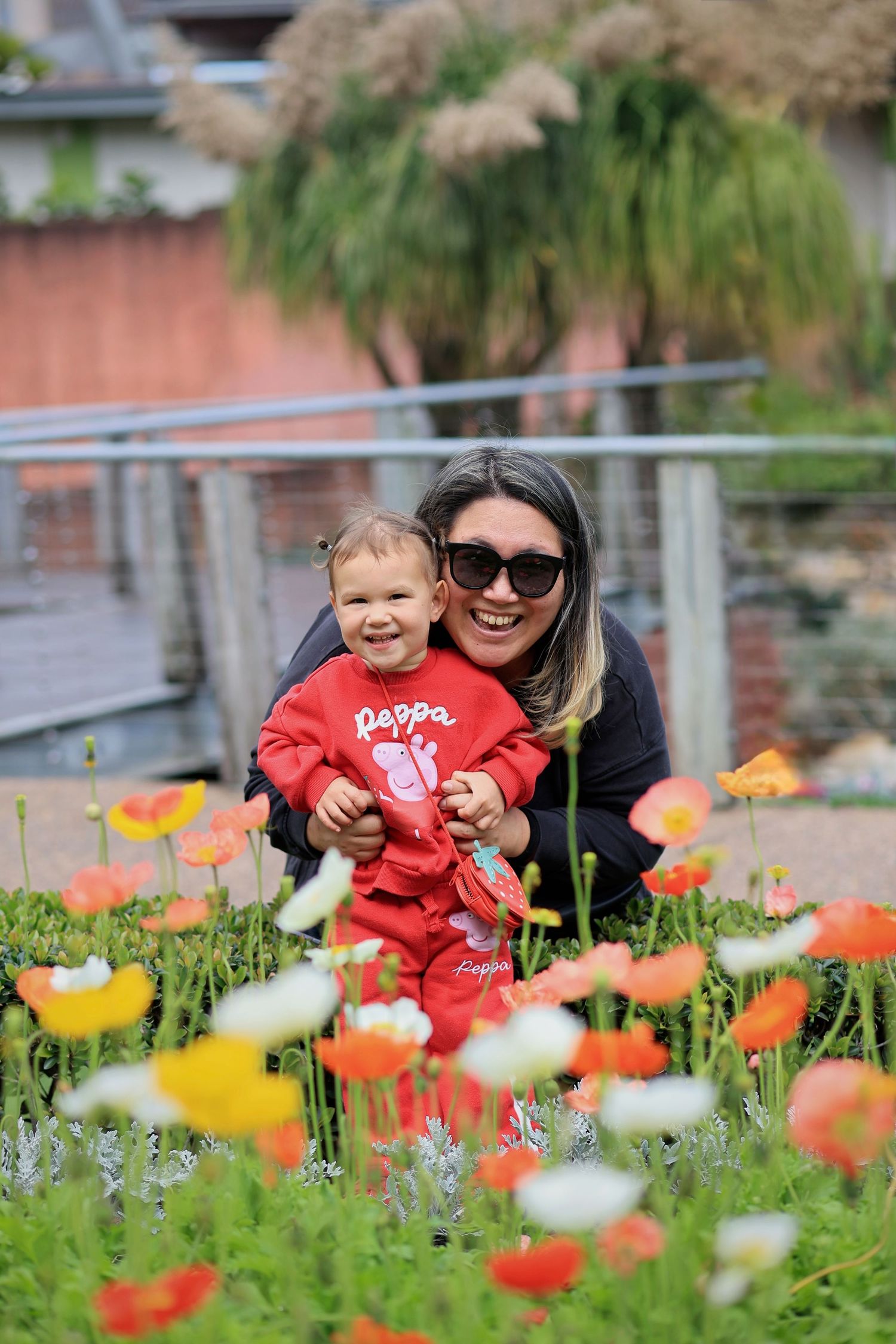 A smiling woman with sunglasses crouches with a baby in red Peppa Pig outfit among colorful poppy flowers in a garden.