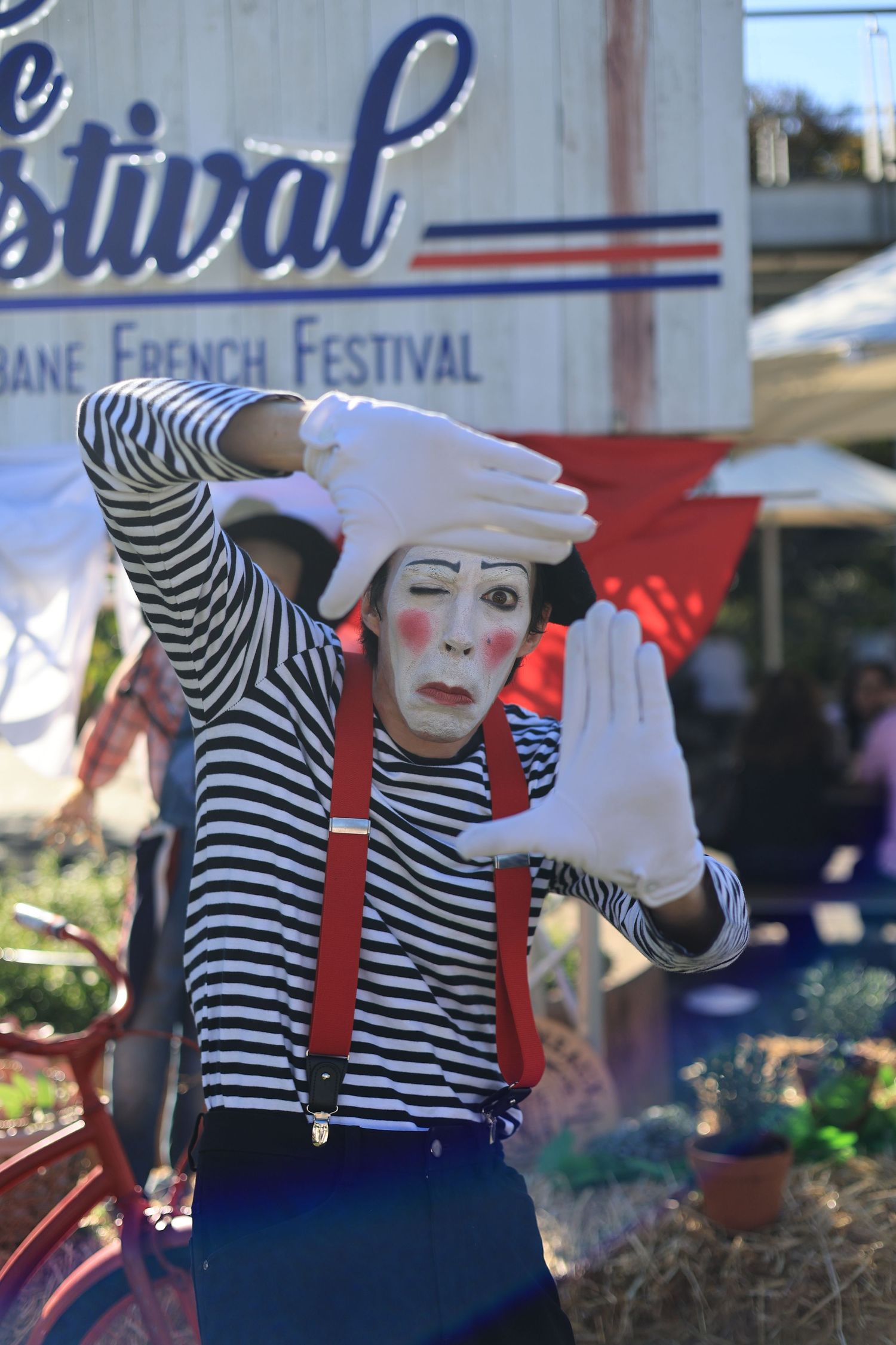 A mime performer in striped shirt, red suspenders, and white face paint frames hands at Brisbane French Festival.