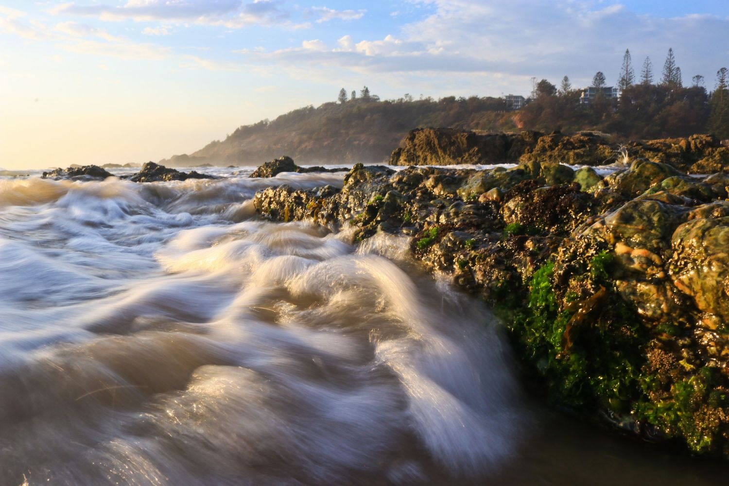 Ocean waves crashing over moss-covered rocks at golden sunset on rocky coastline.