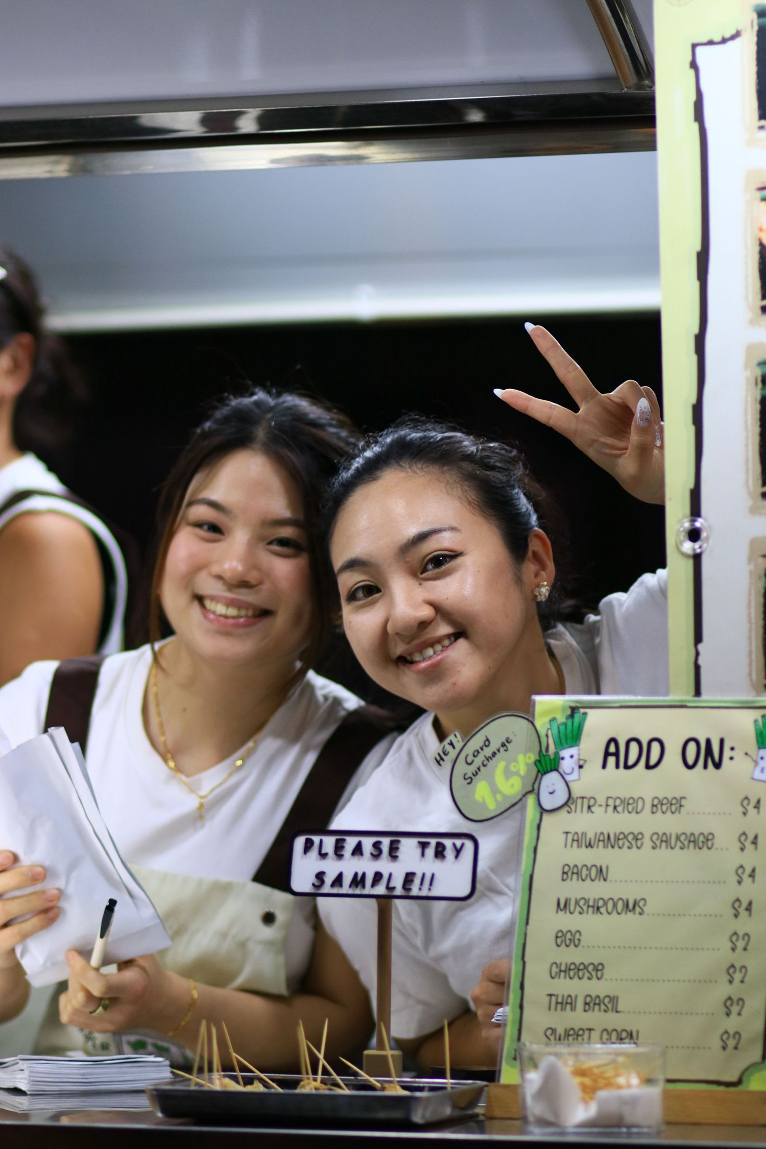 Two smiling young women at a food stall with a please try sample sign and an add-on menu board behind them.