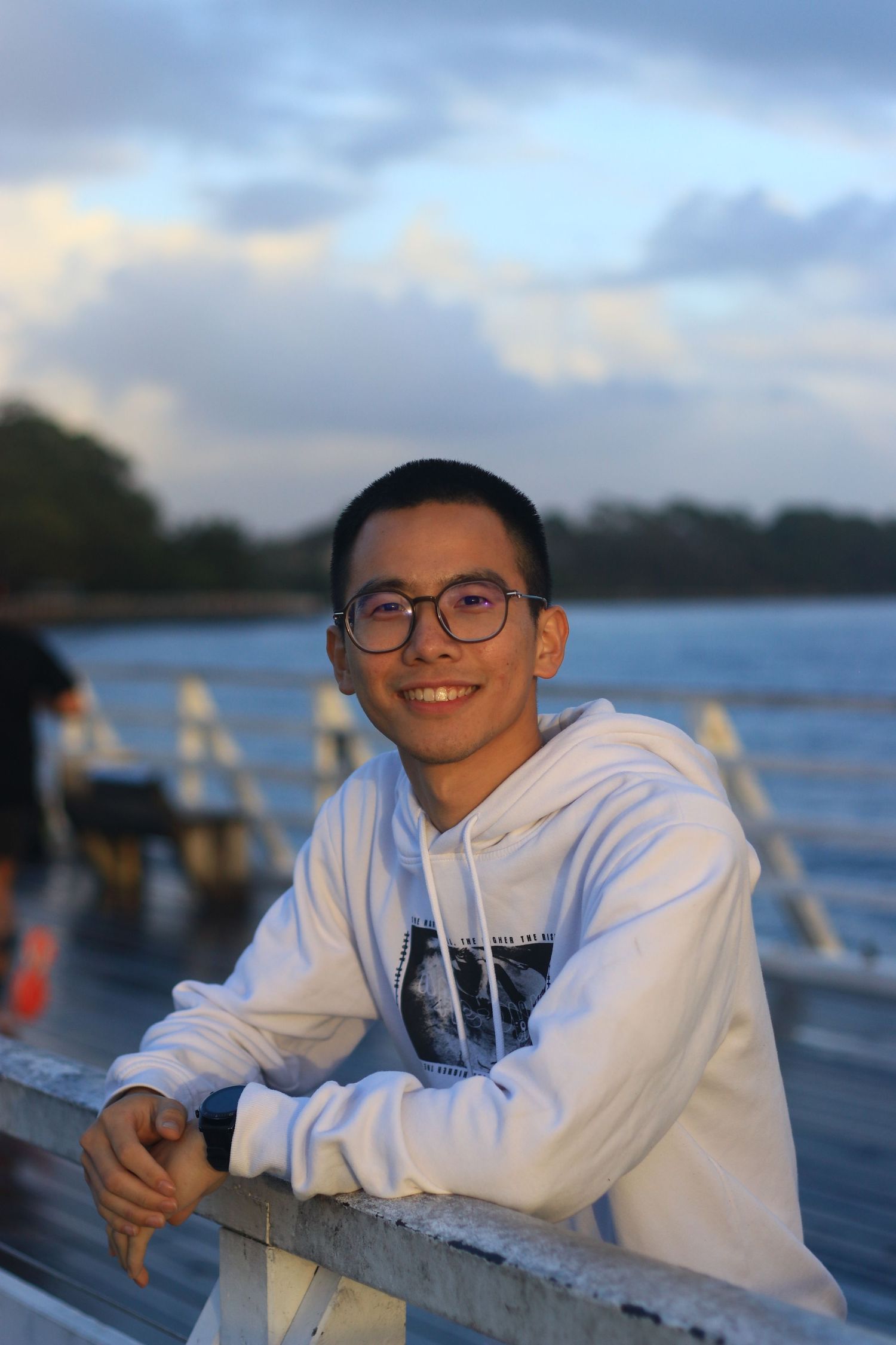 Young Asian man with glasses in white hoodie smiling while leaning on railing near waterfront.