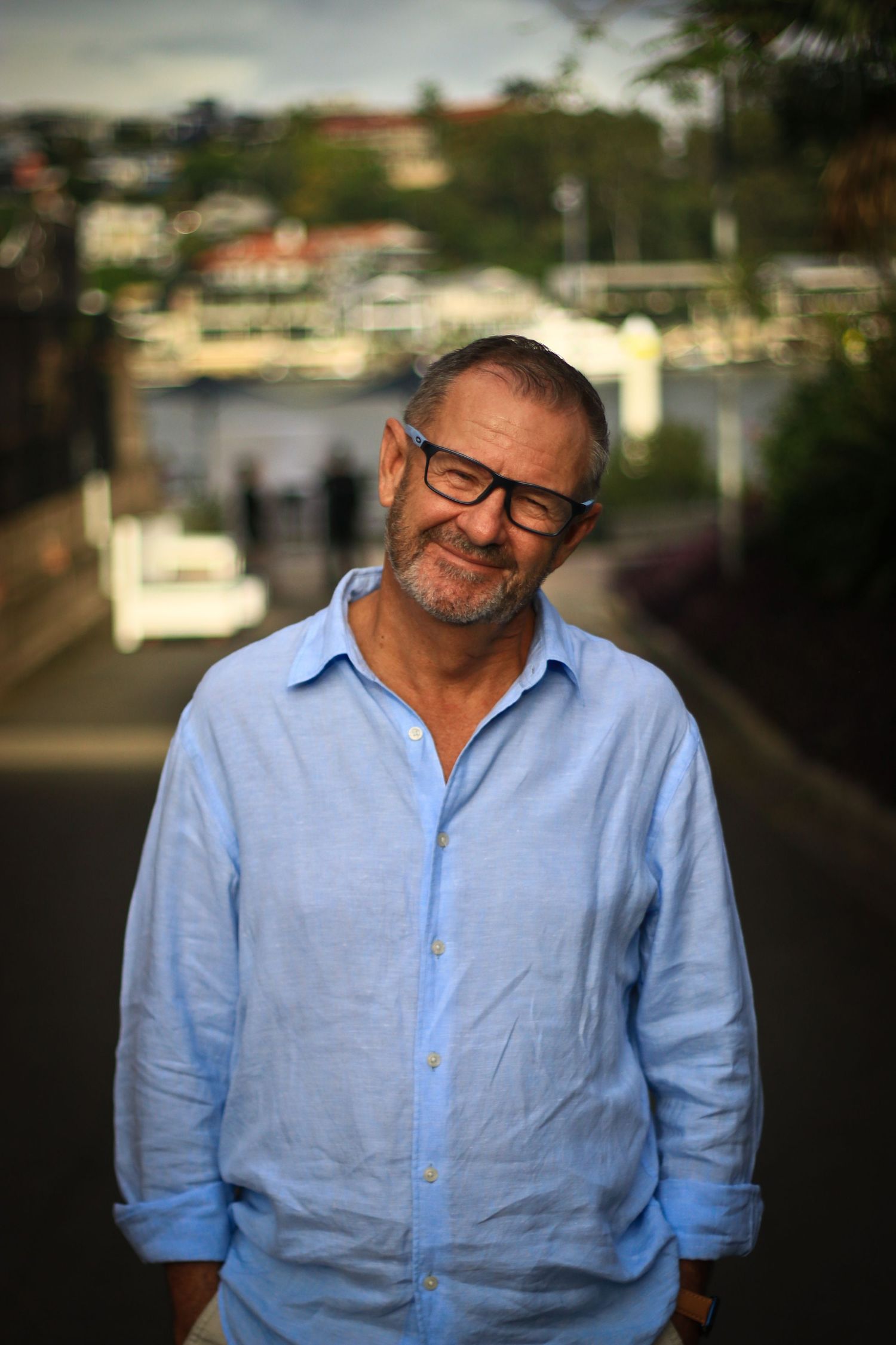 A smiling middle-aged man with glasses and a beard wearing a light blue linen shirt outdoors near a waterway.