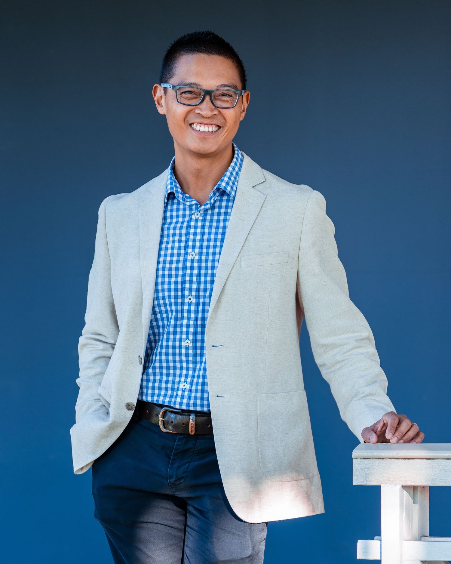Smiling Asian man in light blazer and blue checkered shirt posing against blue background.