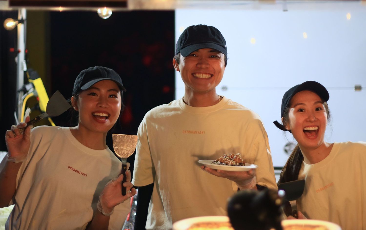 Three smiling food stall workers in matching beige shirts and caps holding food items at a night market.