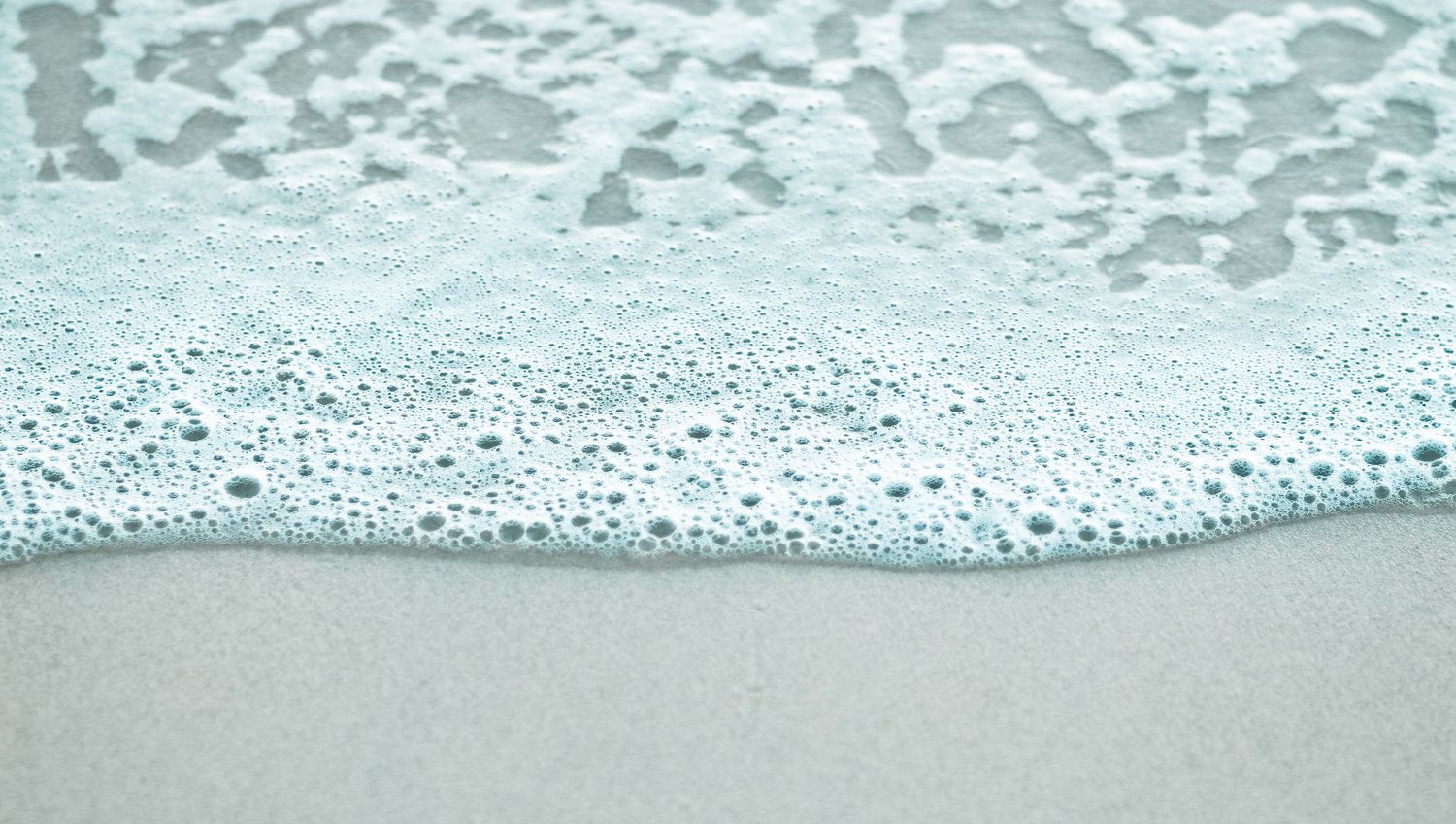 Gentle ocean wave washing over smooth sandy beach with white sea foam.
