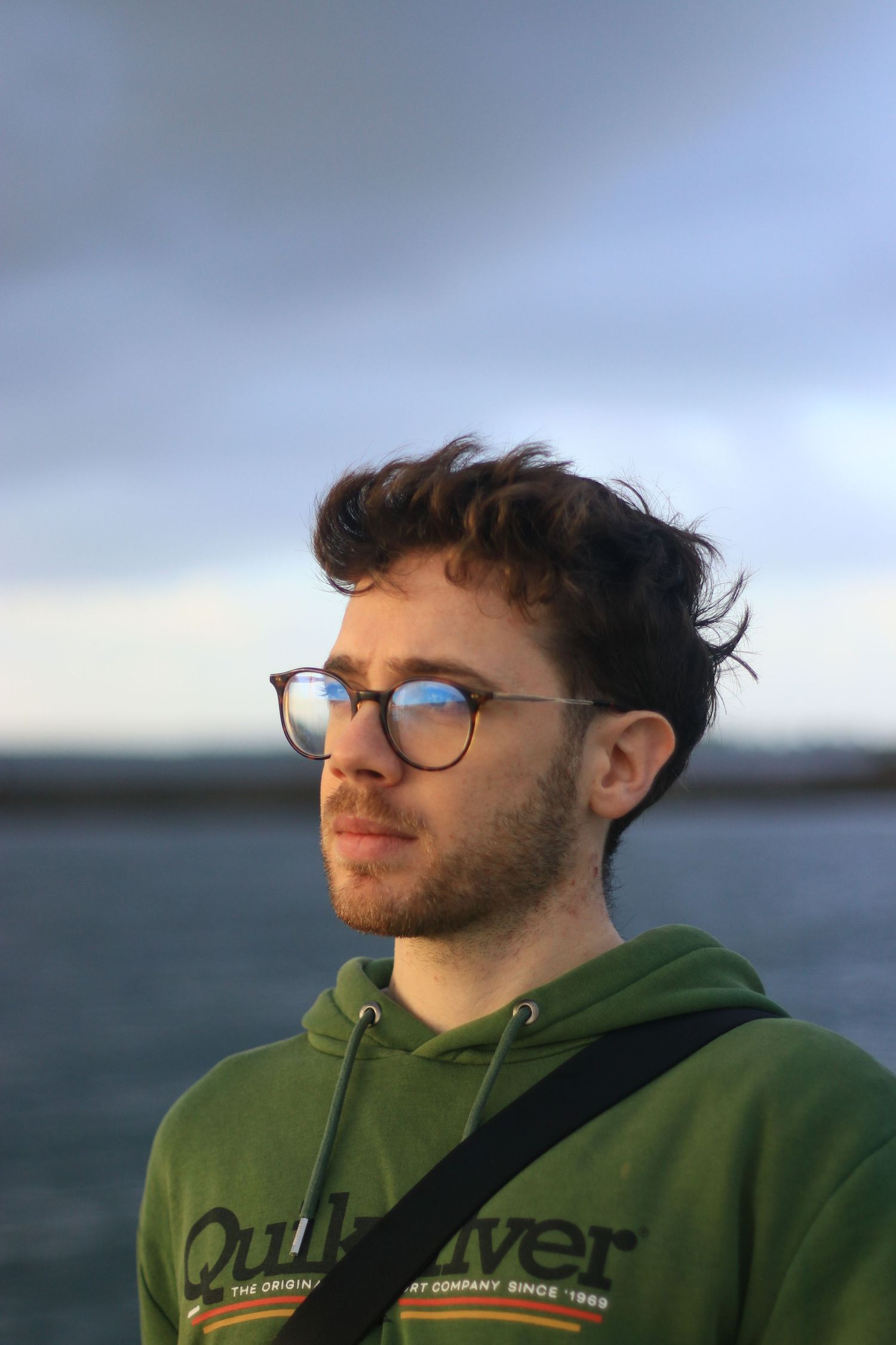 A young man with curly hair and round glasses wearing a green Quiksilver hoodie outdoors near water.