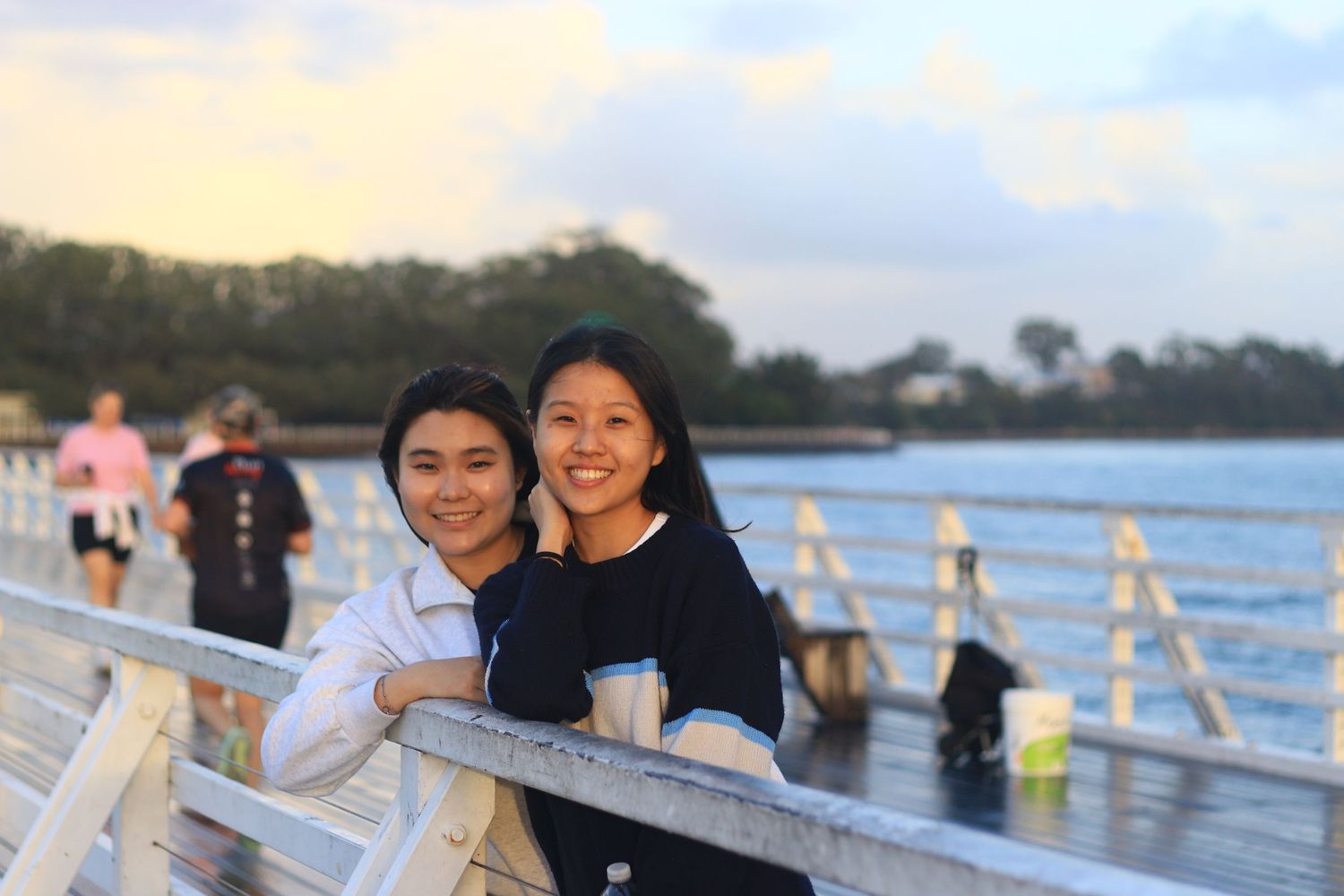 Two young women smiling together on a waterfront pier at sunset with trees in background.